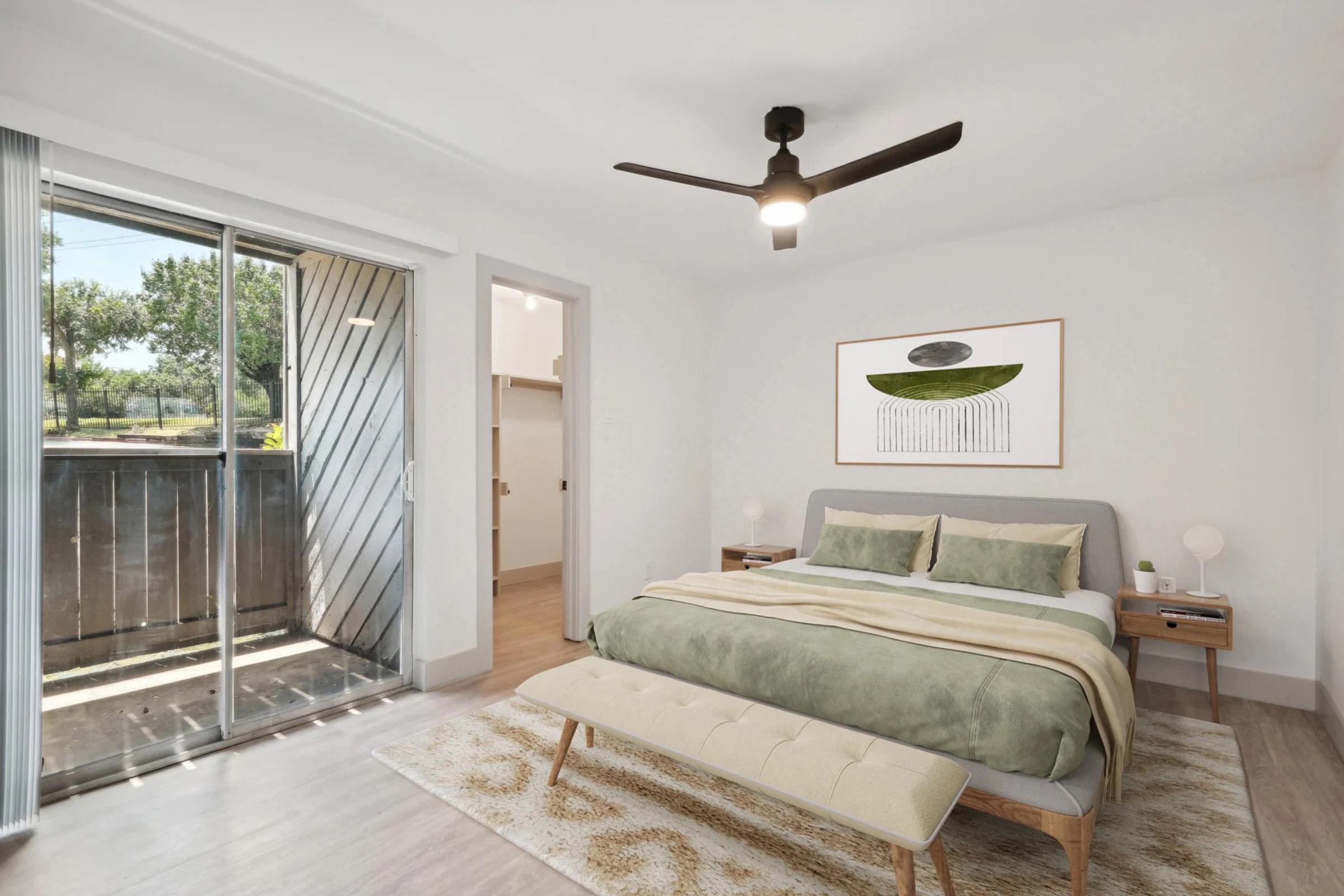 A bright, modern bedroom featuring a queen-sized bed with green and beige bedding, a wooden bench at the foot, and a stylish ceiling fan. There are two bedside tables with lamps, a large window with sliding doors leading to a balcony, and a piece of abstract art on the wall, all set against a light-colored backdrop.