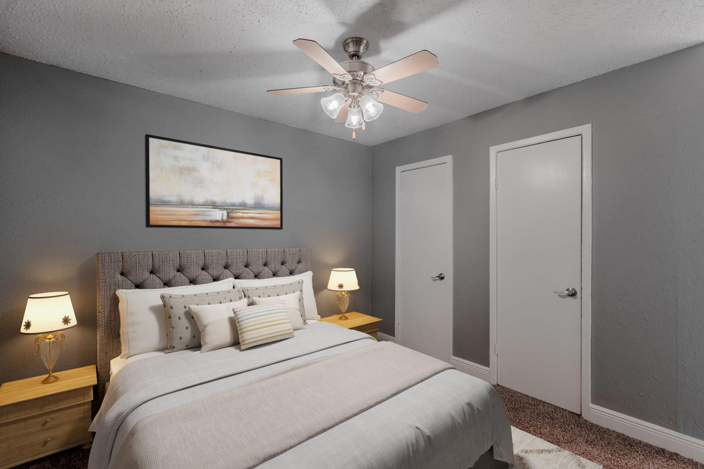 A cozy bedroom featuring a tufted gray headboard with white bedding and decorative pillows. Two bedside tables with lamps flank the bed, and a painting adorns the wall above. The room has a ceiling fan, light gray walls, and two white doors, creating a calm and inviting atmosphere.