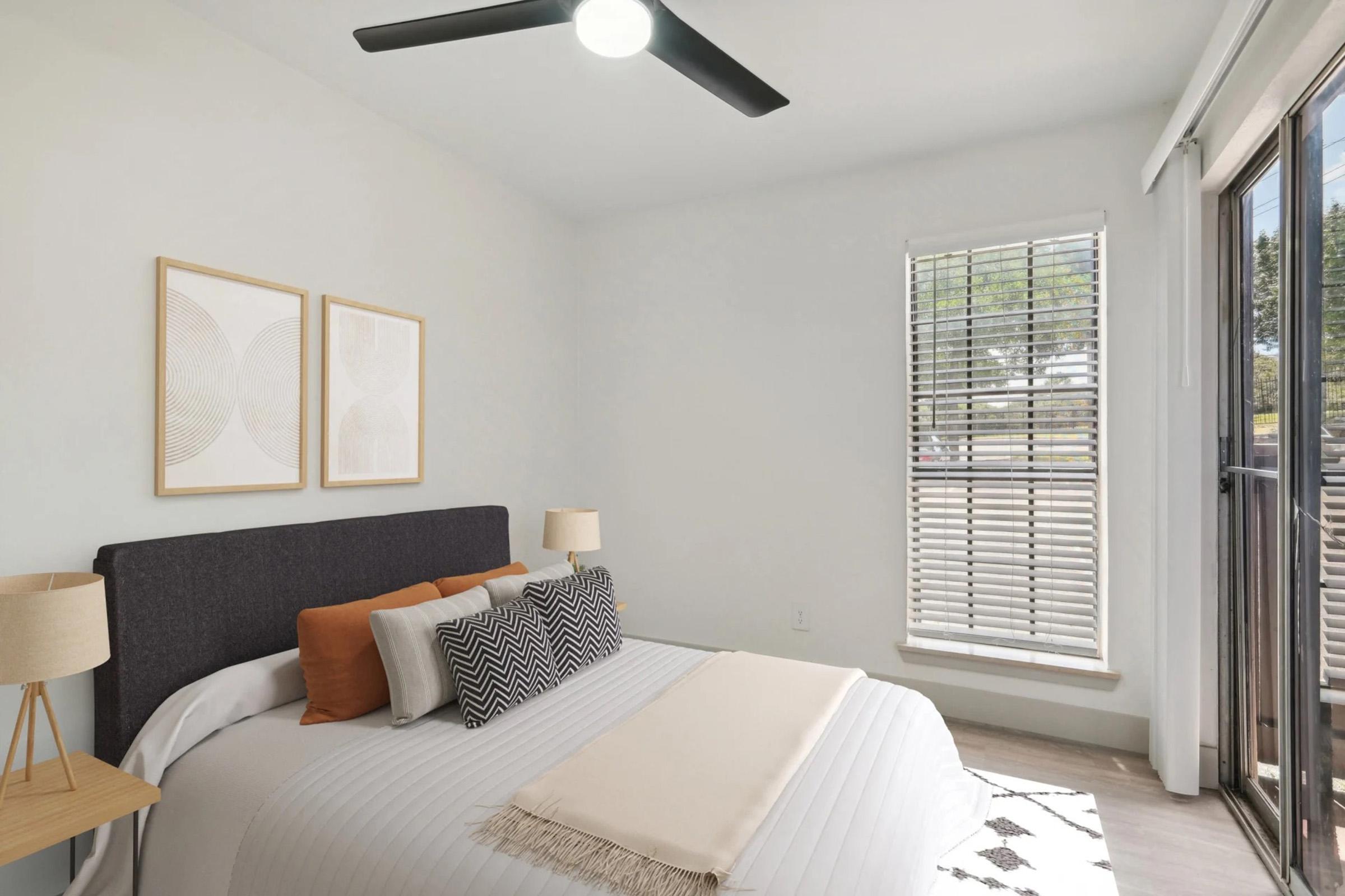 A well-lit bedroom featuring a king-sized bed with neutral bedding and decorative pillows. There are two bedside lamps on matching tables, and the walls are adorned with circular art pieces. A large window with blinds allows natural light to fill the space, complementing the modern design.