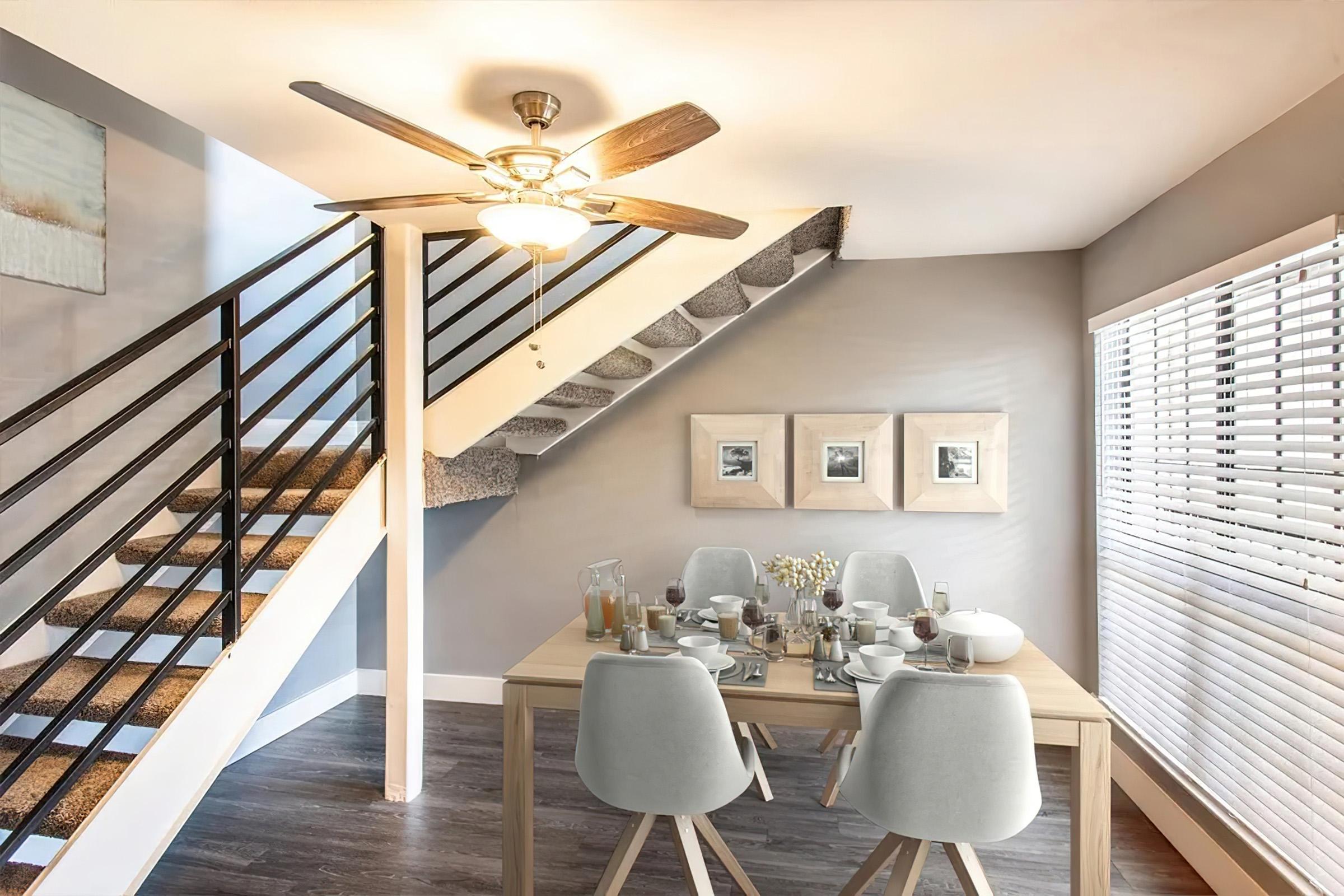 A modern dining area featuring a wooden table set for a meal, with elegant dishes and drinks. A staircase with a sleek railing is visible in the background, alongside large windows with blinds that let in natural light. The walls are painted in soft colors, and decorative frames adorn the wall.