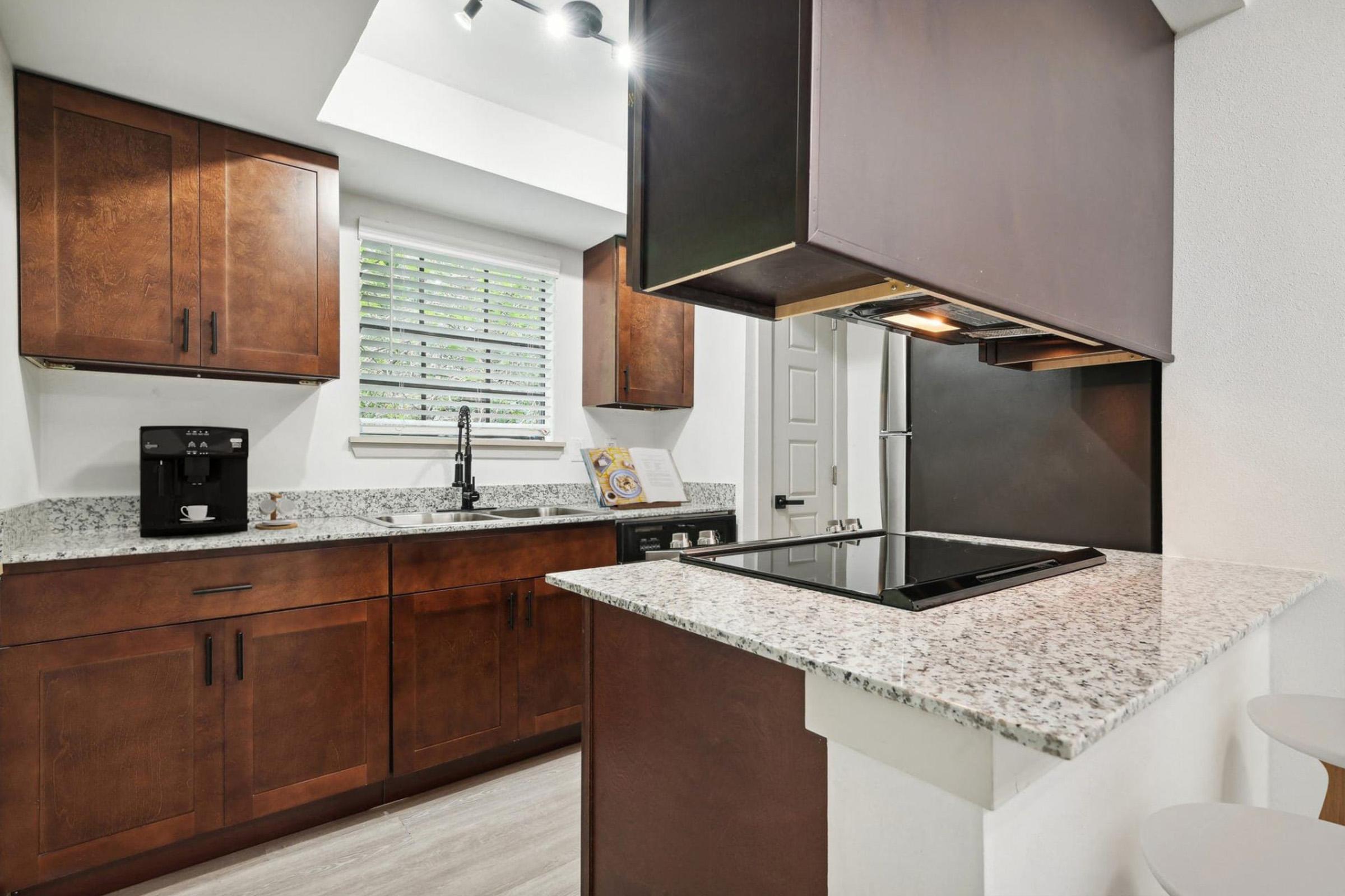 Modern kitchen featuring rich wooden cabinets, a granite countertop, and an island with a stovetop. The space includes a window with blinds allowing natural light, a coffee maker on the counter, and sleek appliances, creating a stylish and functional cooking area.