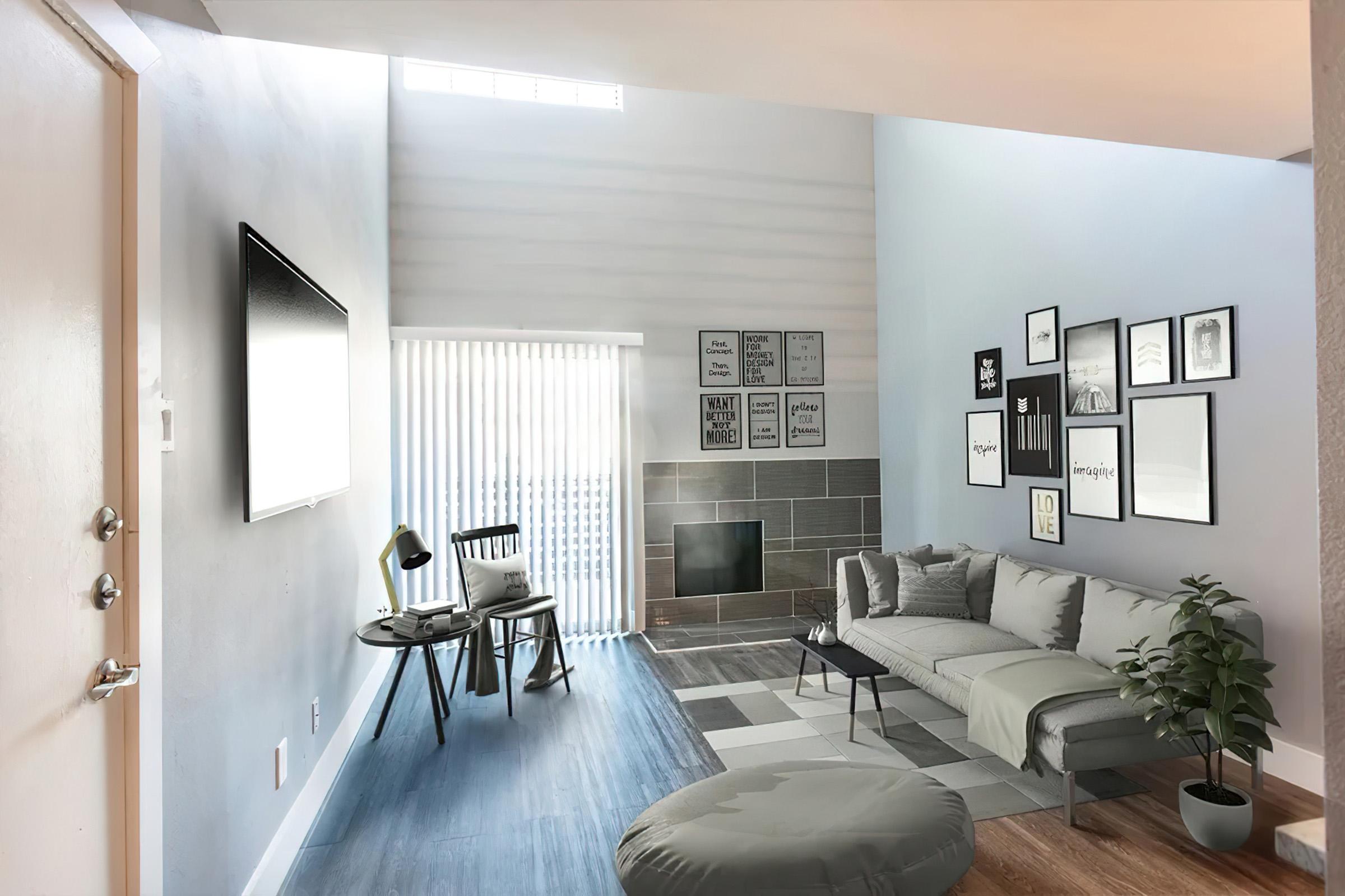 A modern living room featuring a gray sectional sofa, round ottoman, and a wall-mounted TV. Large windows with blinds allow natural light in, illuminating framed art on the walls. A small table and chair sit beside the sofa, and a potted plant adds a touch of greenery to the stylish, minimalist decor.