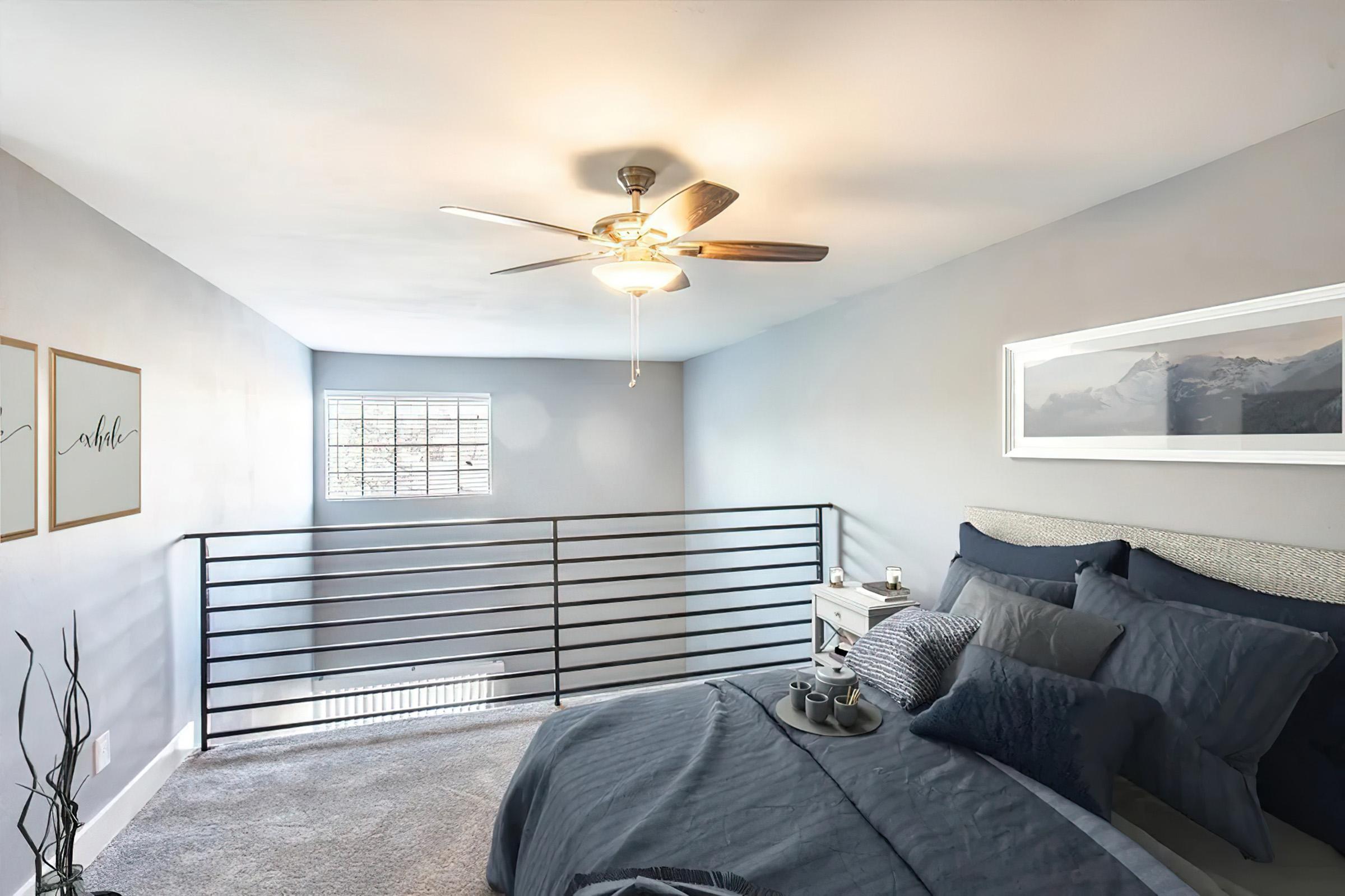 A cozy bedroom featuring a ceiling fan, light gray walls, and soft lighting. The space includes a neatly made bed with dark blue bedding, a small nightstand with decorative items, and a modern railing overlooking a lower level, enhancing the open feel of the room. A large window allows natural light to fill the space.