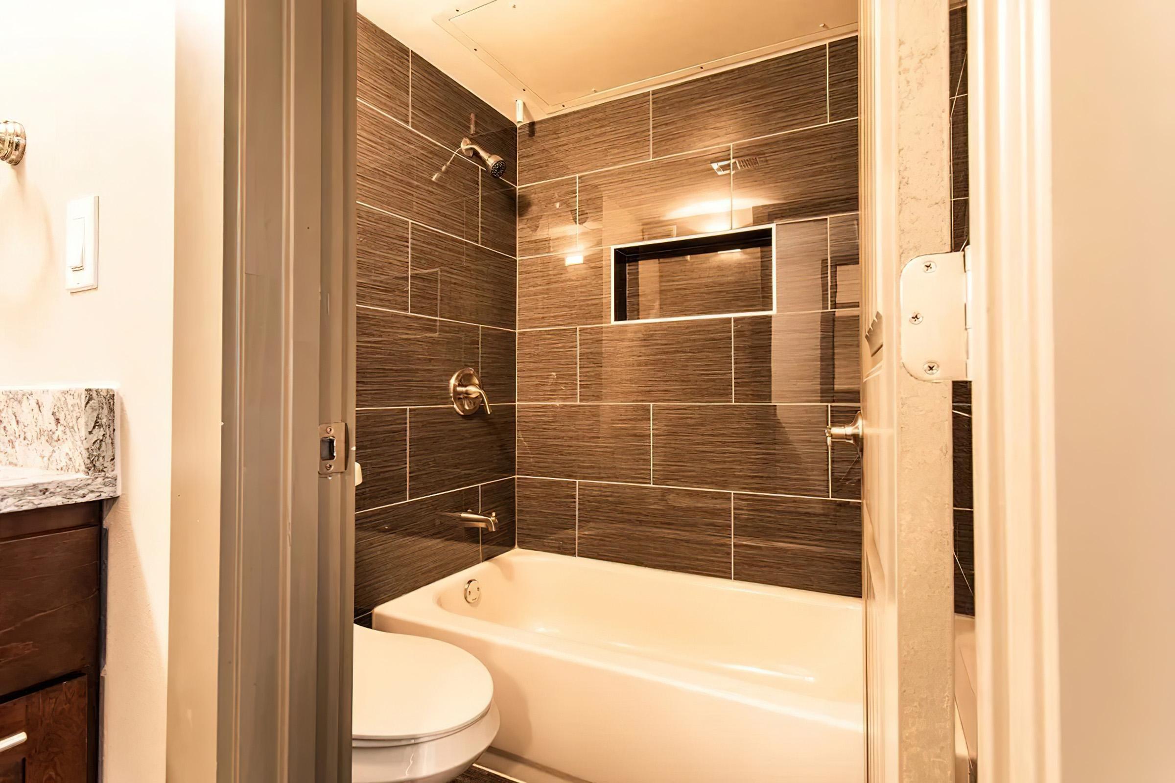 A modern bathroom featuring dark tiled walls, a white bathtub, and a sleek shower. There are two wall-mounted lights and a glass window above the bathtub. The bathroom door is partially open, revealing a glimpse of a wooden vanity with a granite countertop.
