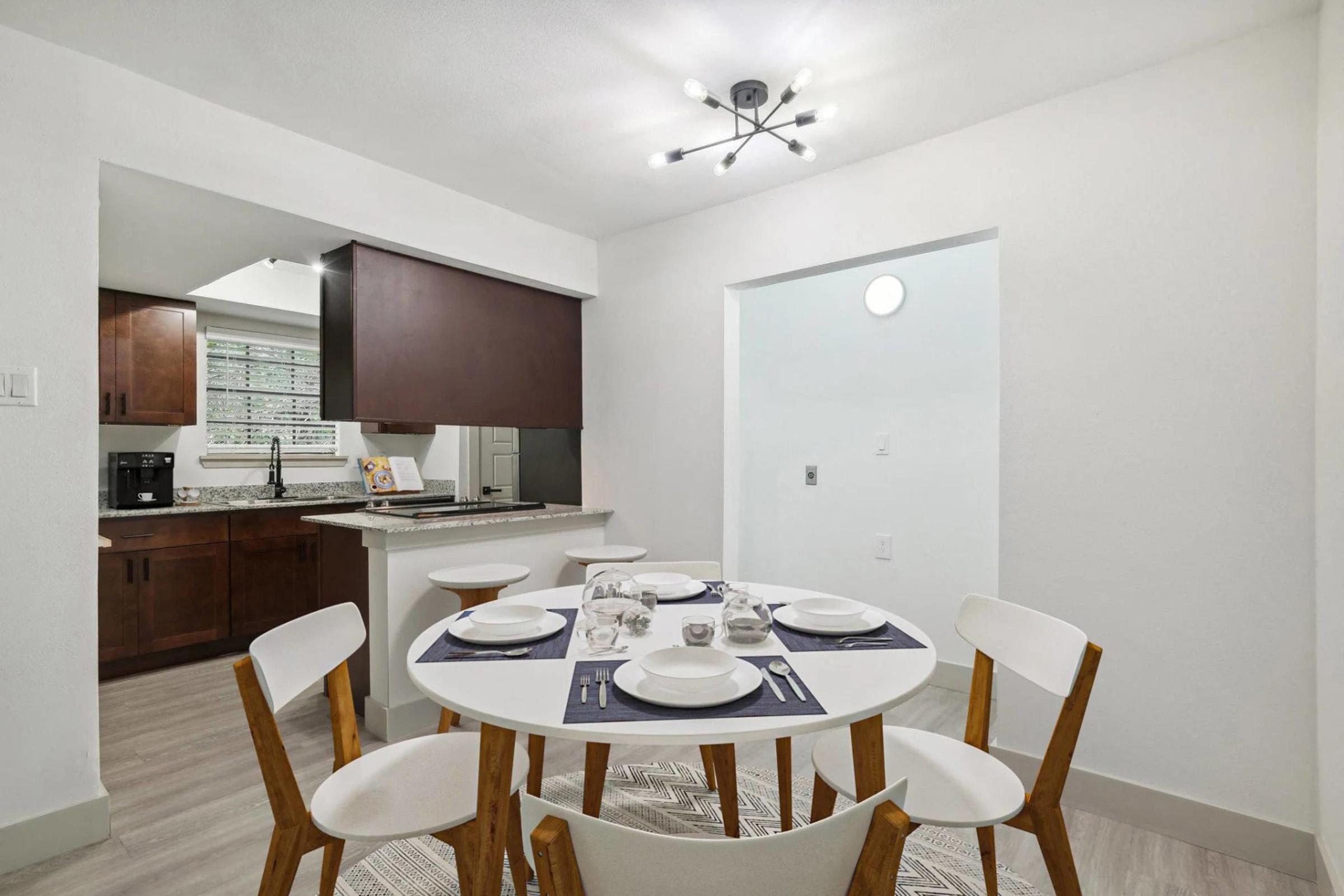 A modern dining area featuring a round white table set with plates and utensils, surrounded by white chairs. In the background, a kitchen area with dark wood cabinets and a granite counter is visible, along with natural light coming in from a nearby window. The walls are painted in light colors, creating a bright ambiance.