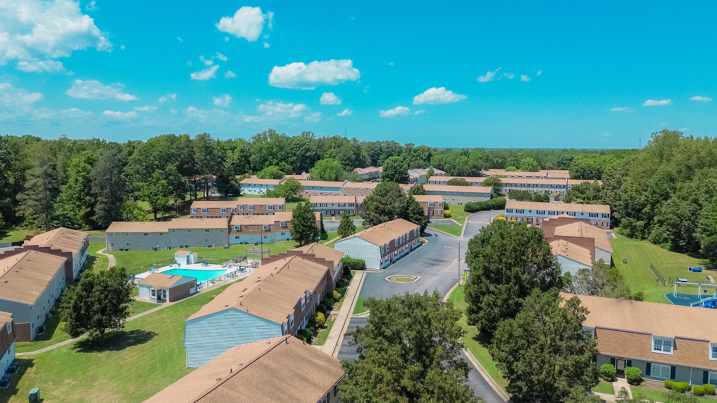 Aerial view of a residential neighborhood featuring multiple two-story buildings with brown roofs, surrounded by green trees and grassy areas. A swimming pool is visible in the center, and a well-maintained road weaves through the complex, with clear blue skies overhead and a few clouds.
