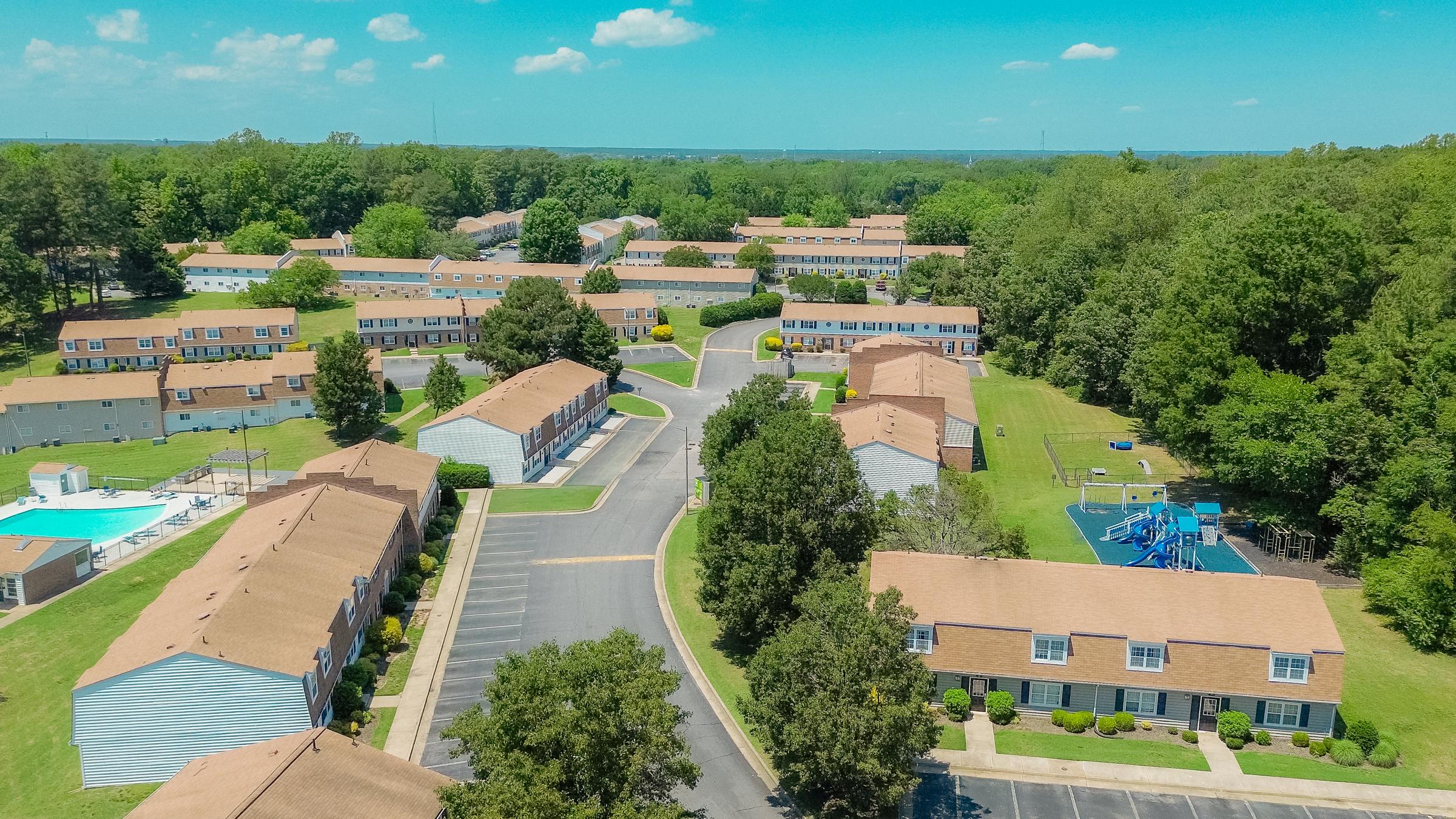 Aerial view of a residential community featuring multiple two-story buildings with brown roofs, surrounded by green lawns and trees. A swimming pool is visible on the left, and a playground can be seen in the background. The layout includes winding driveways and parking areas, creating a spacious atmosphere.