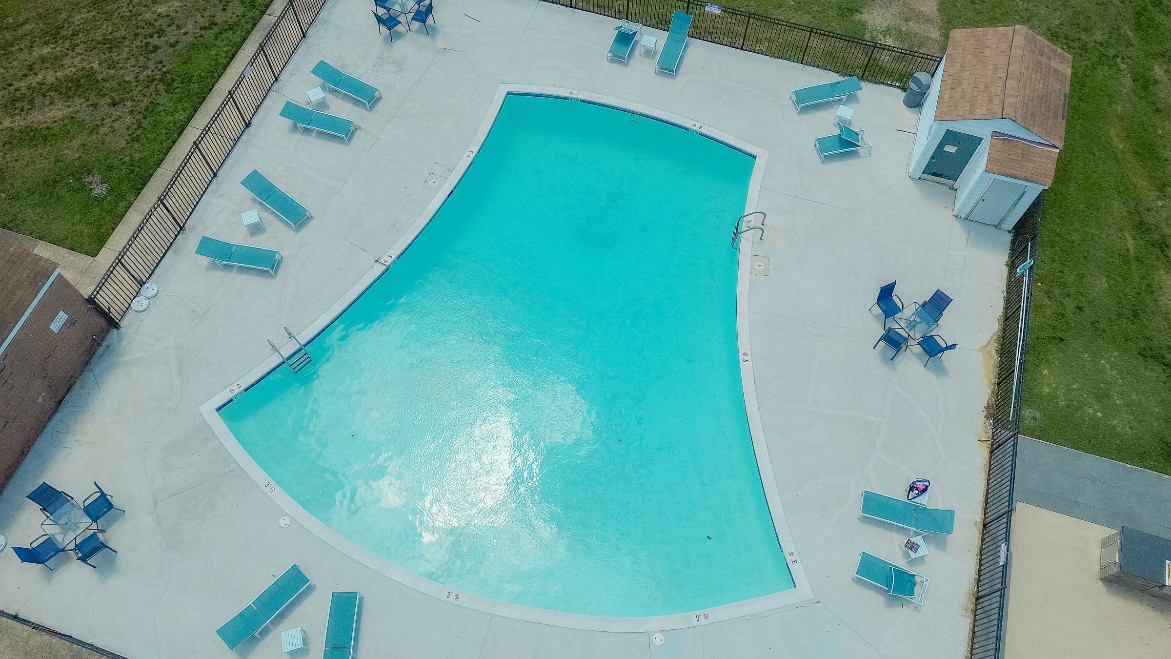 Aerial view of a swimming pool with a unique shape, surrounded by lounge chairs and patio furniture. Nearby, there is a small shed and a grassy area. The water is clear and inviting, reflecting sunlight.