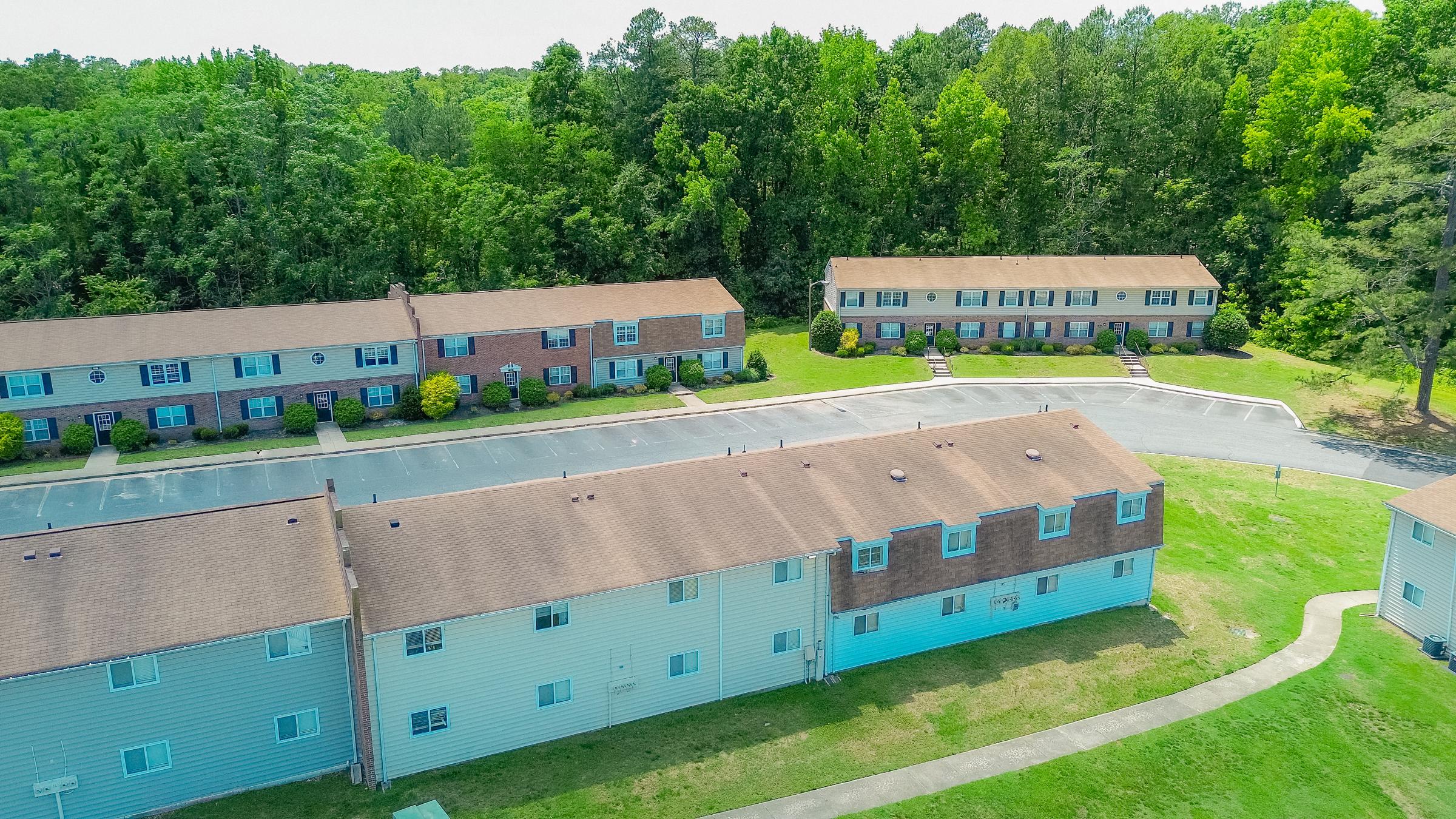 Aerial view of a residential complex with two rows of two-story apartment buildings surrounded by greenery. The buildings have brown and blue siding, with a paved parking area in front. Lush trees and foliage are visible in the background, indicating a suburban setting.