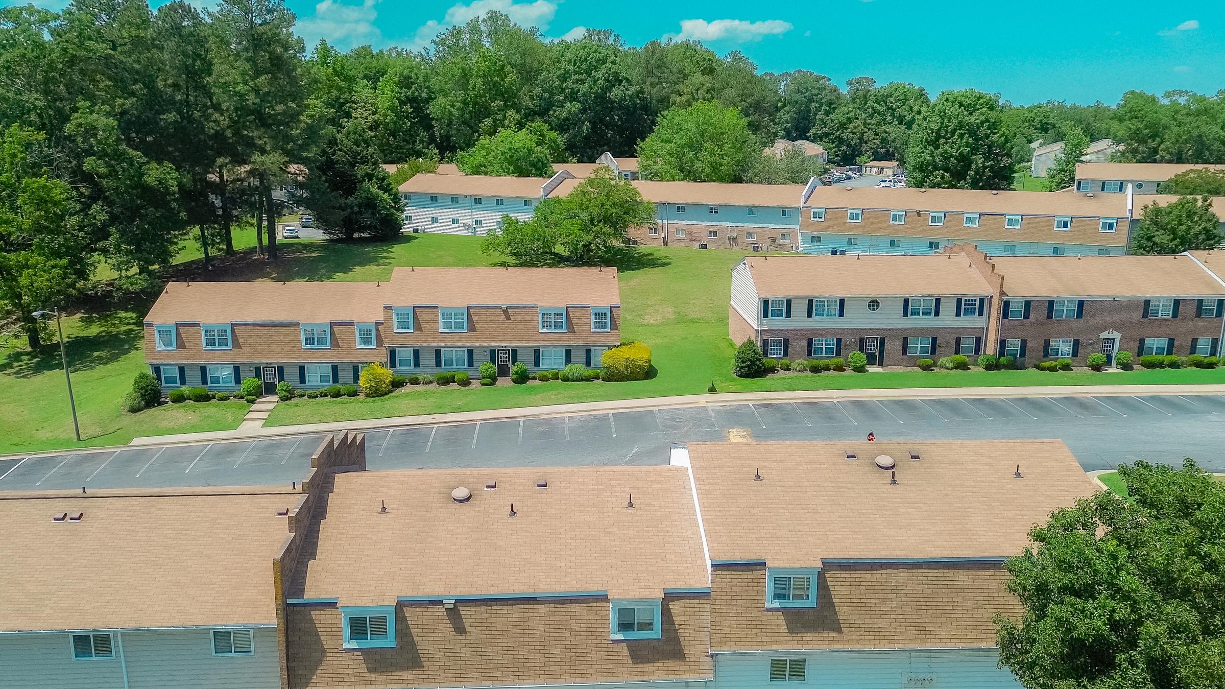 Aerial view of a residential area featuring two rows of townhouses with brown roofs. Lush green lawns and trees surround the buildings, and additional multi-story apartments are visible in the background. The scene is set on a clear, sunny day with a bright blue sky.