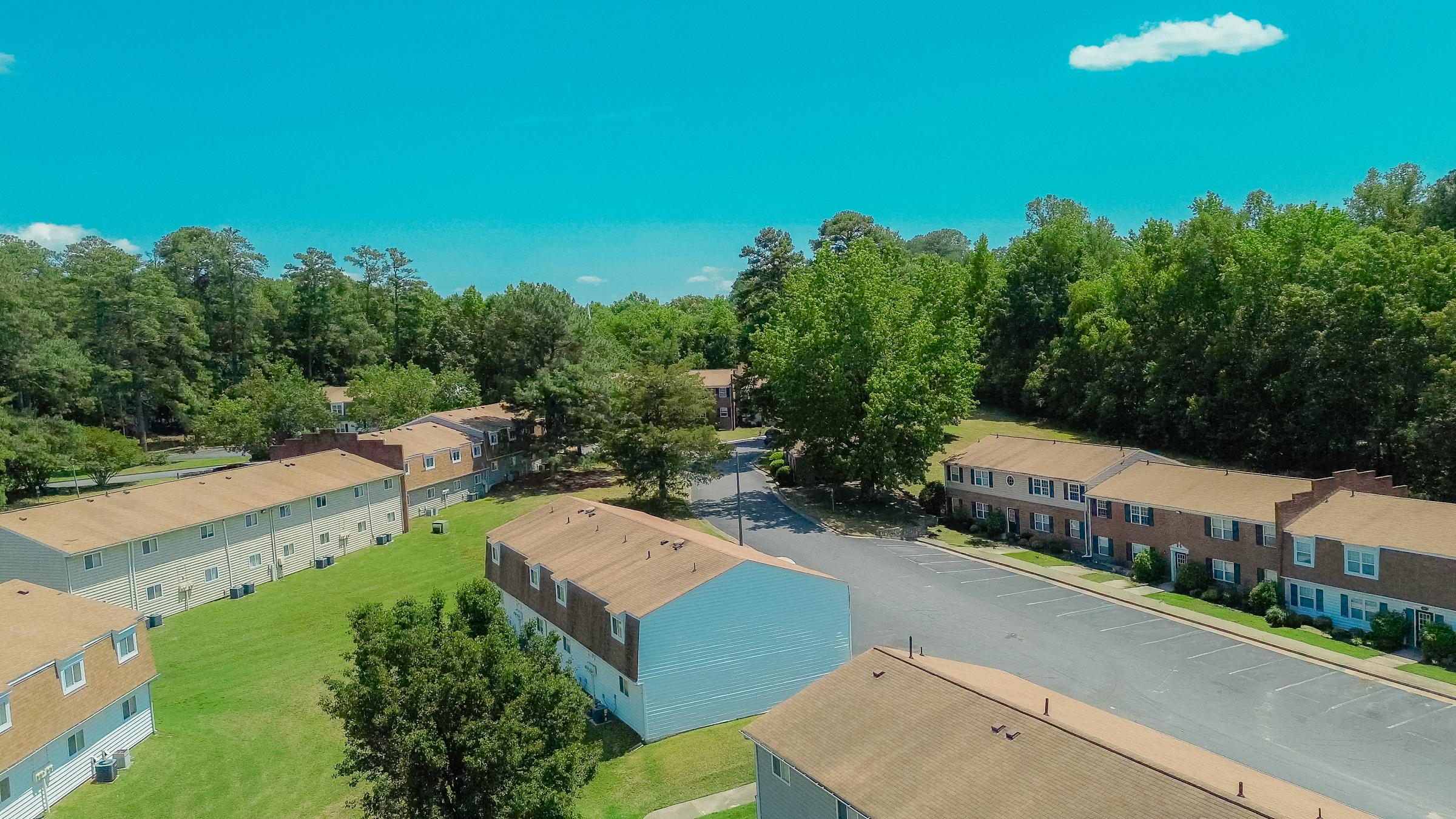 Aerial view of a residential neighborhood featuring several two-story buildings with brown roofs. Green lawns and trees surround the buildings, creating a scenic landscape. The sky is clear blue with a few fluffy white clouds. A winding road runs through the area, leading to more greenery in the background.