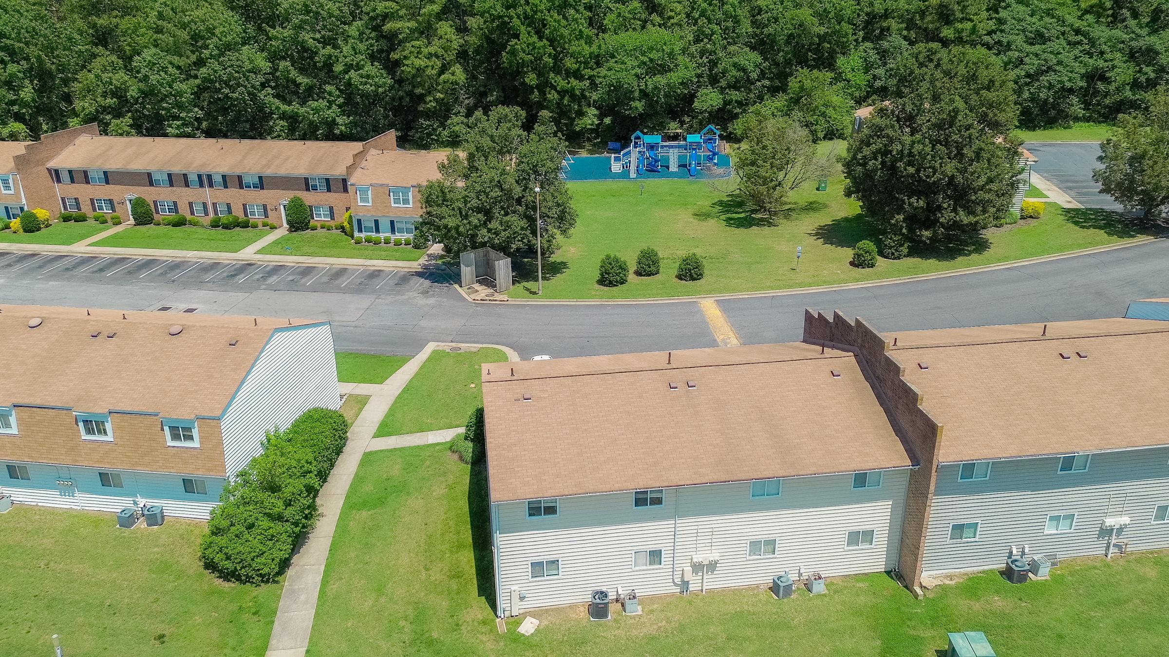 Aerial view of a residential area featuring two buildings with brown roofs. One building has a light-colored exterior, while the other is a two-story structure with a similar color. In the background, there is a grassy area with playground equipment and trees lining the edge of the scene. Parking spaces are visible along the street.