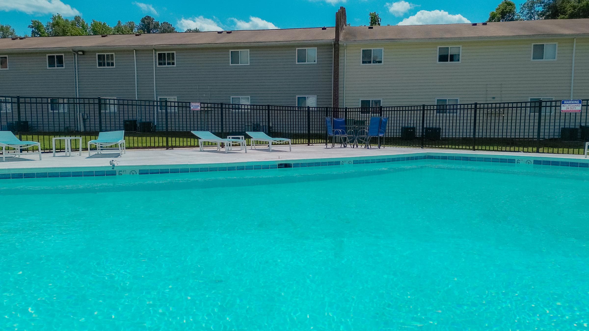 A clear blue swimming pool surrounded by lounge chairs and a fence. In the background, there are several residential apartment buildings under a bright blue sky with a few clouds.