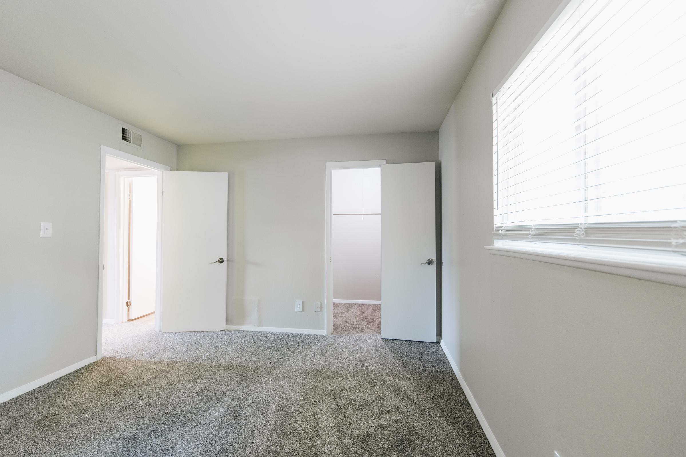 A spacious, empty room with gray walls and carpet. It features two closed doors, one leading to a closet and the other to a hallway. A window on the far wall provides natural light, with white blinds partially covering it. The room has a clean and modern feel, suitable for various uses.