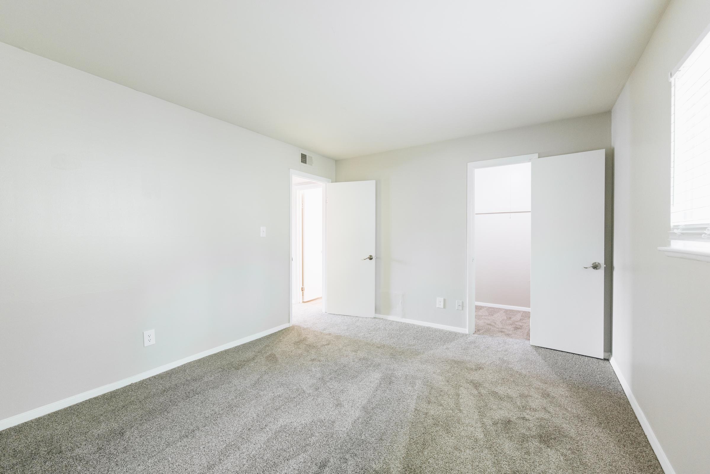 A spacious, empty room with light gray walls and freshly carpeted flooring. Two doors are visible: one leading to a closet and the other to an adjacent room. Natural light streams in through a small window, illuminating the minimalistic space. Ideal for a bedroom or office setup.