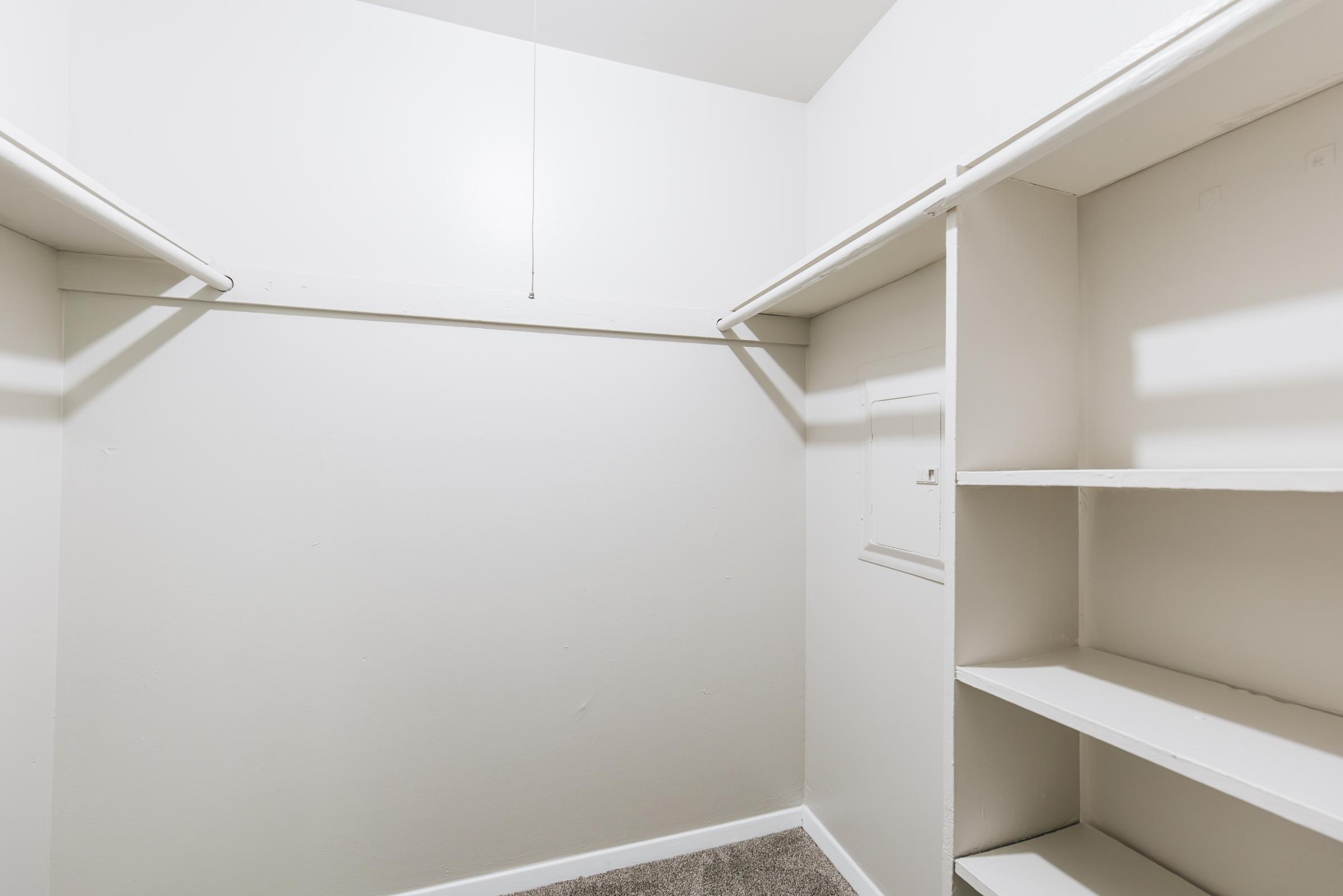 Empty closet space with white walls and a carpeted floor. Features two hanging rods for clothing and shelves on the right side for storage. The lighting is bright, and there is a small panel on the wall, indicating a simple and functional design.