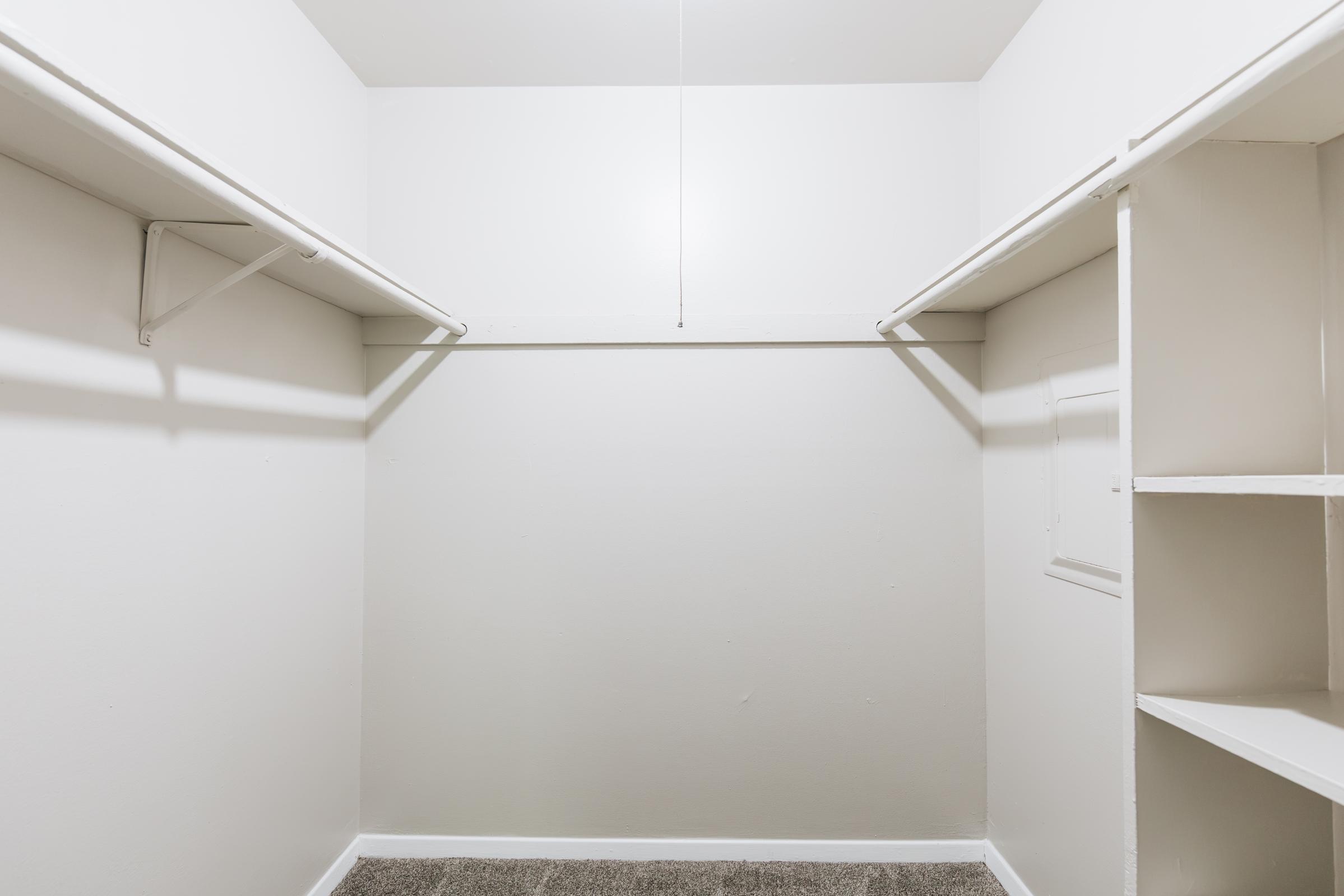 A clean, empty closet with white walls and shelving. The closet features two horizontal rods for hanging clothes and a small shelf on one side, with a light fixture hanging from the ceiling. The floor is carpeted in a neutral color.