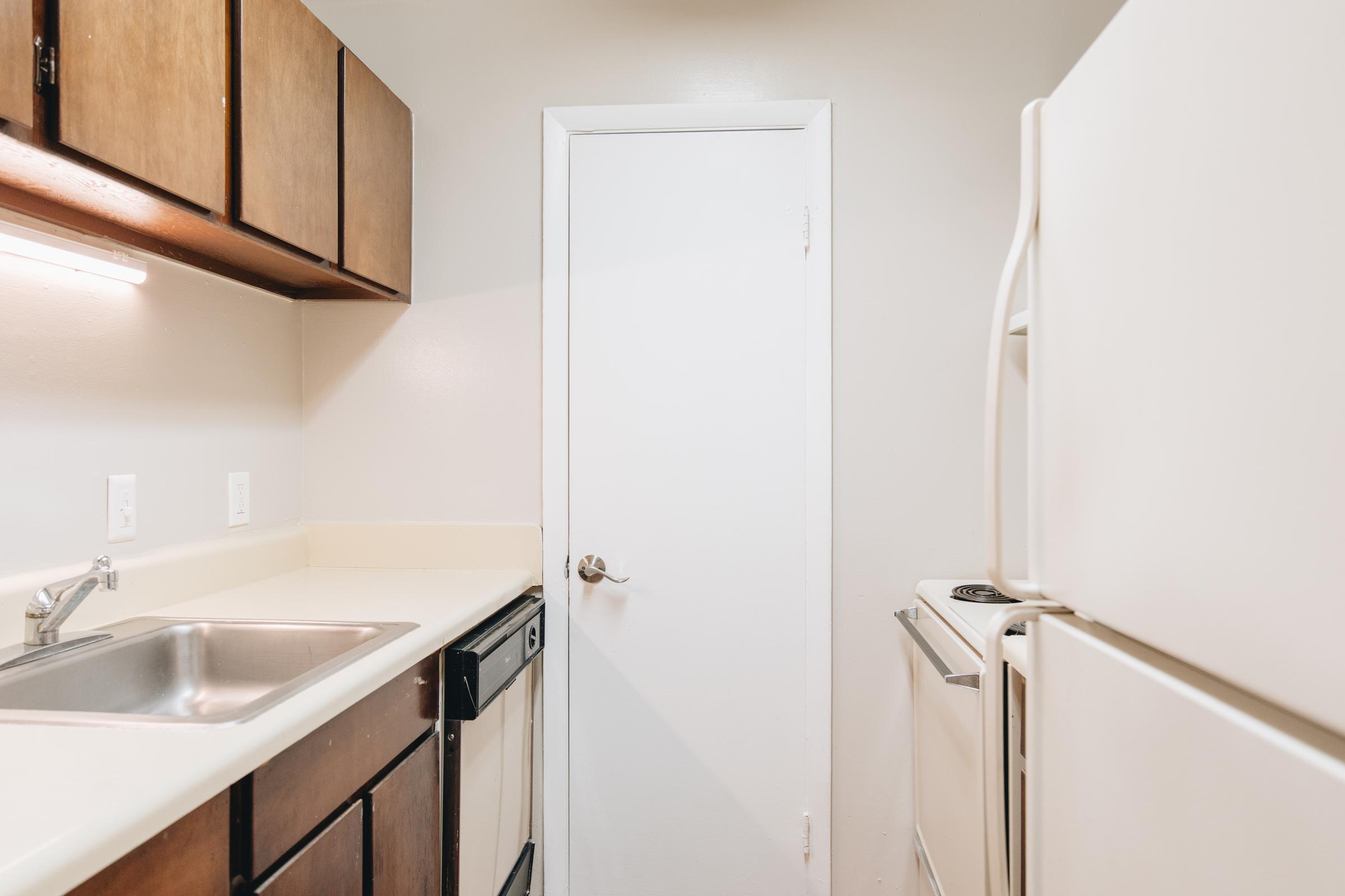 A small kitchen featuring wooden cabinets, a stainless steel sink, a white refrigerator, and a closed door. The countertops are light-colored, and the walls are painted in a neutral tone. The space appears well-lit and organized, suitable for basic cooking needs.