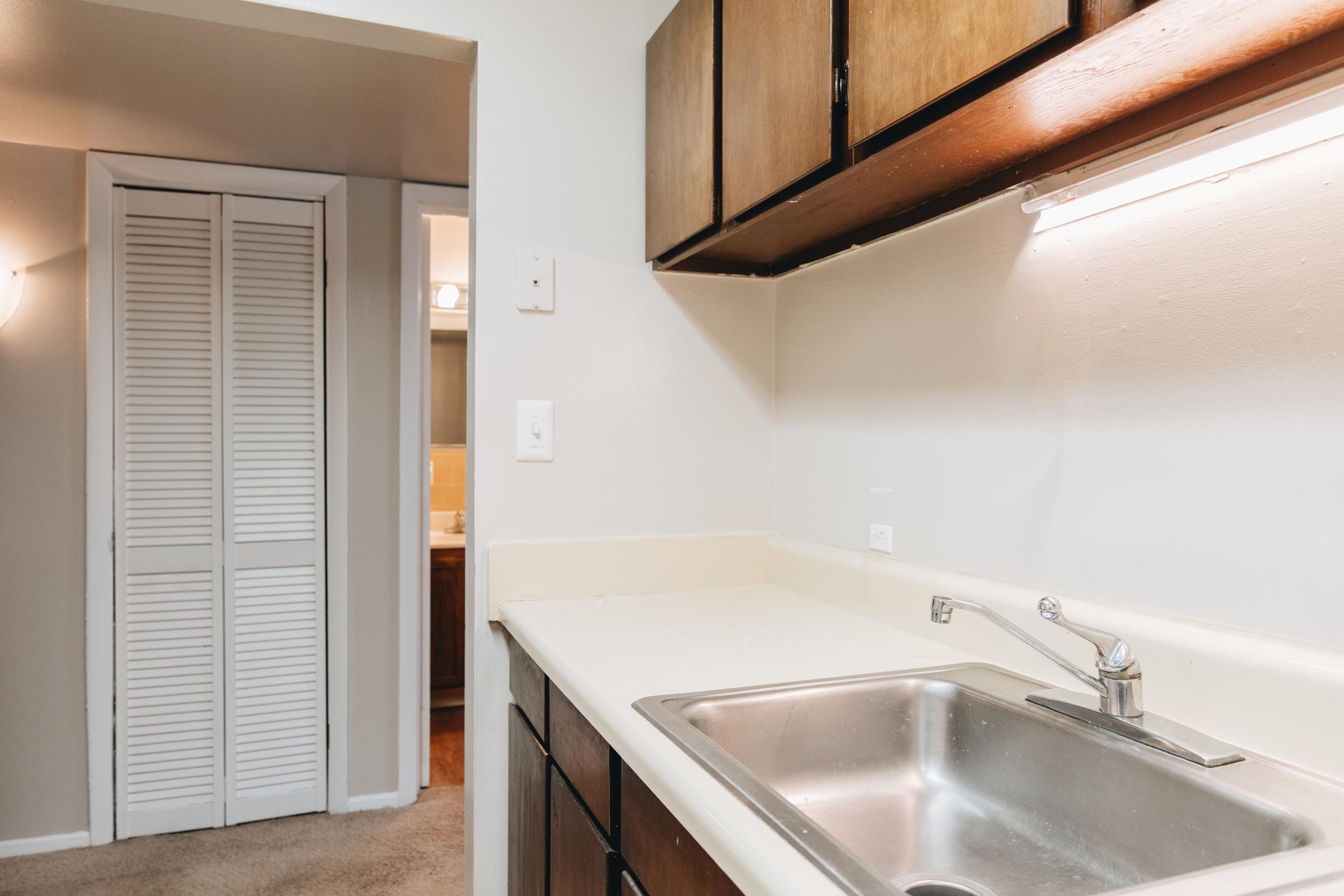 A small kitchen area featuring a stainless steel sink, beige countertop, and dark wooden cabinetry. The background shows a doorway leading to another room, likely a bathroom, with a light-colored wall and a closet with louvered doors.