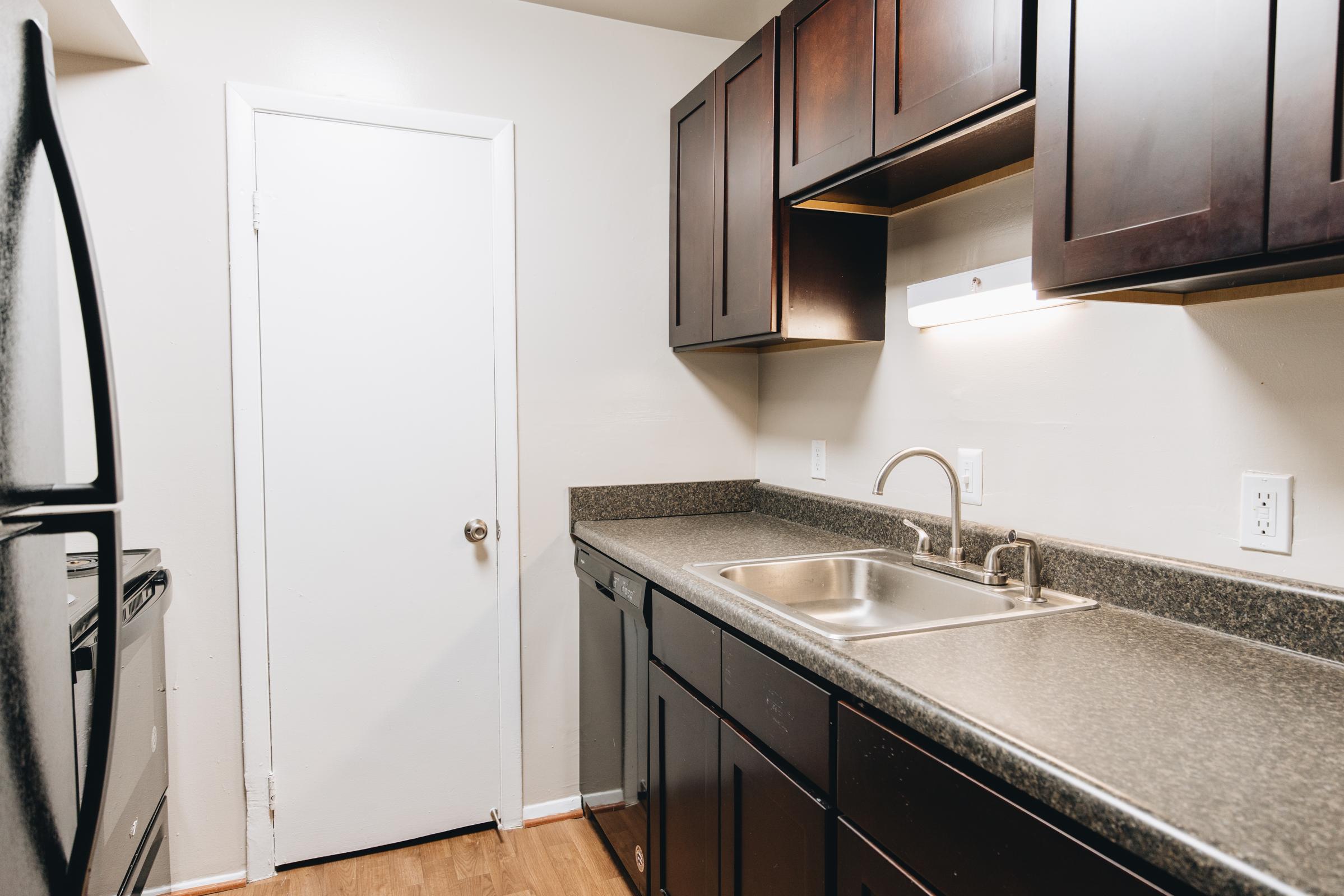 A modern kitchen featuring dark wood cabinets, a stainless steel sink with a faucet, and a countertop. The appliances include a black refrigerator and an oven. The walls are painted a light color, and there is a closed white door on one side. The flooring is wooden, adding warmth to the space.