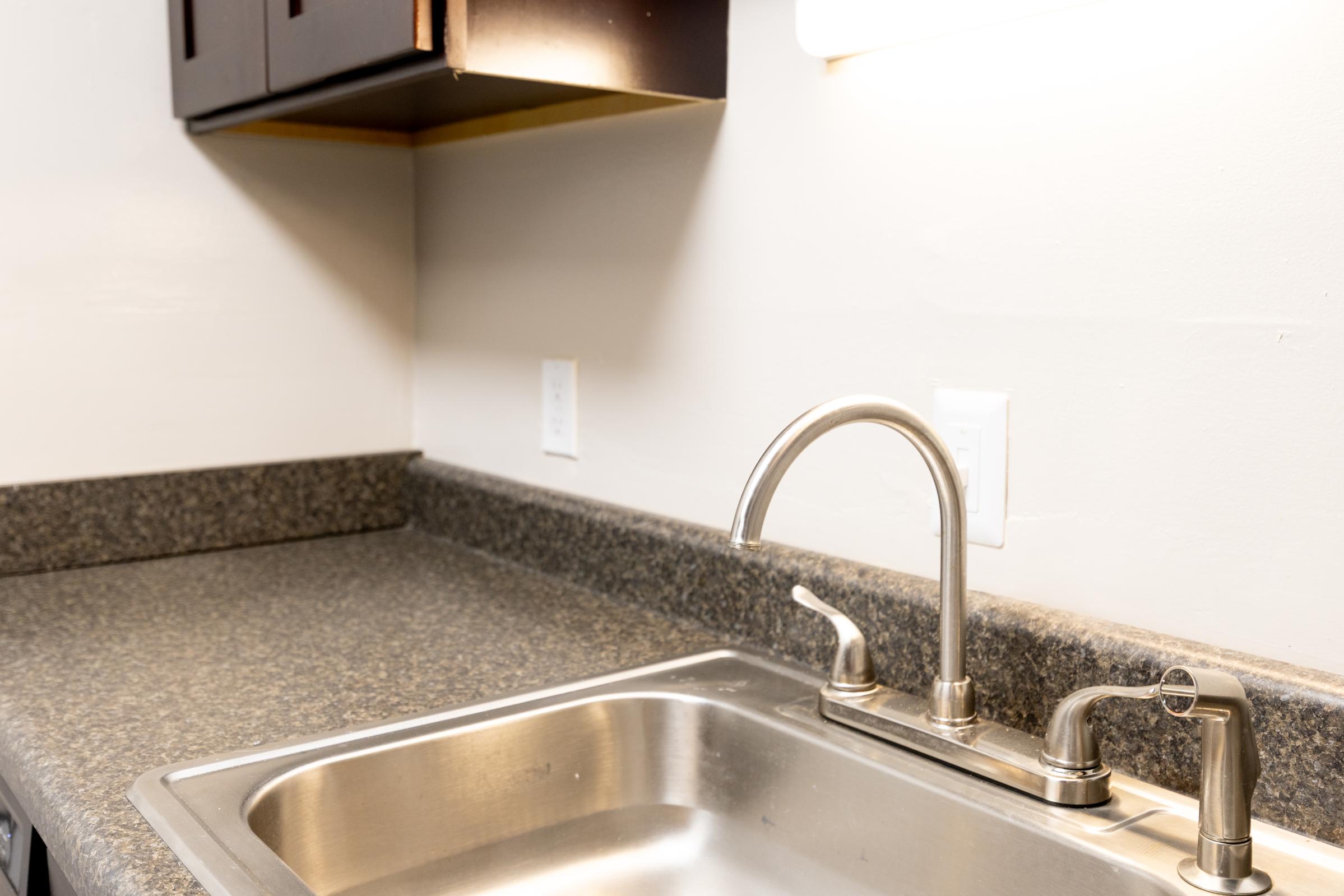 A kitchen sink with a stainless steel basin and a polished metal faucet, set against a granite countertop. Above the countertop, there are upper cabinets. The wall is painted a light color, and there is a light fixture installed above the sink area. The scene is well-lit and modern.