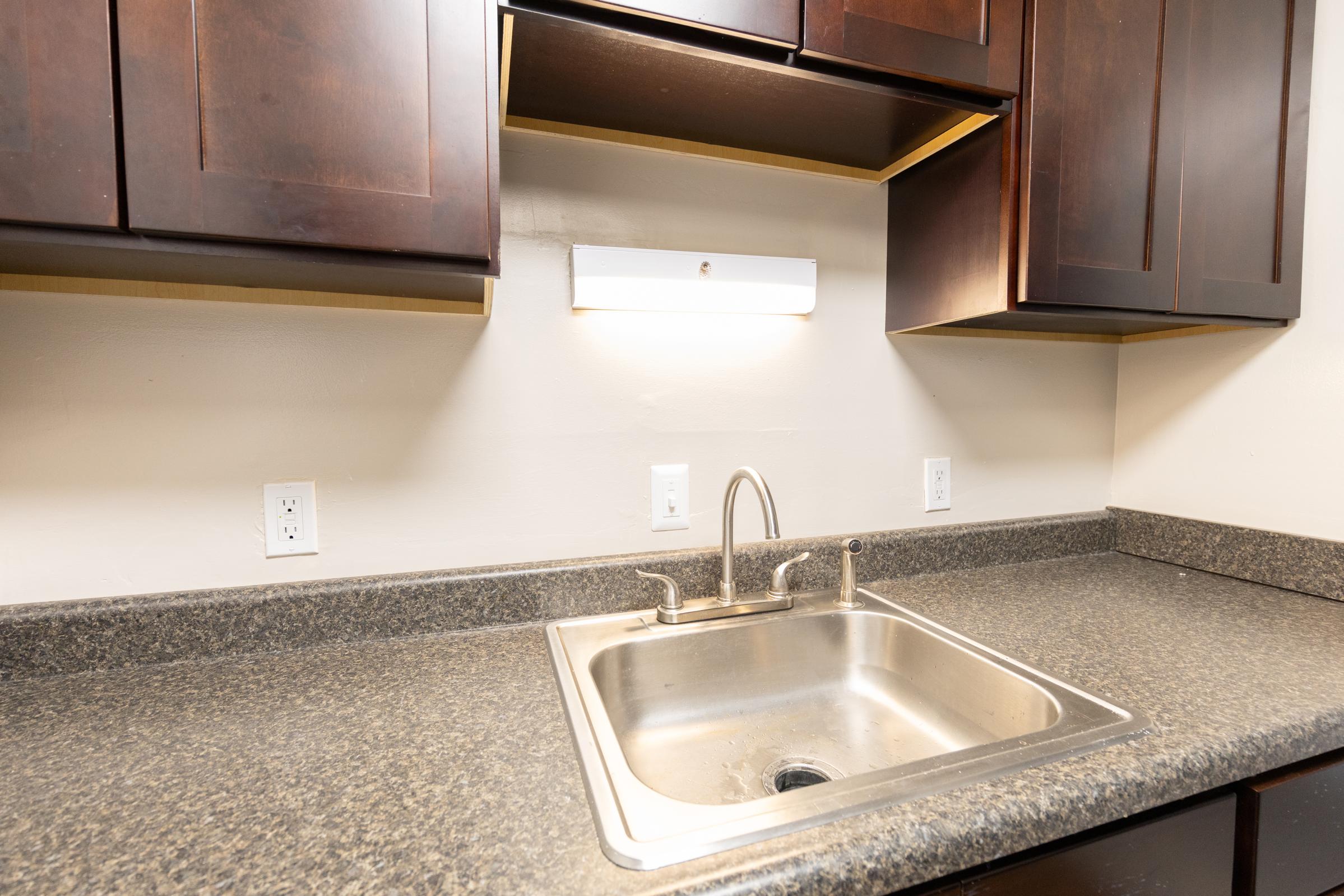 A clean kitchen counter with a stainless steel sink and modern faucet. Above the sink, there is a strip light, and dark wooden cabinets are mounted on the wall. The countertop is dark and smooth, providing a contemporary look to the space. Plug outlets are visible on the wall.
