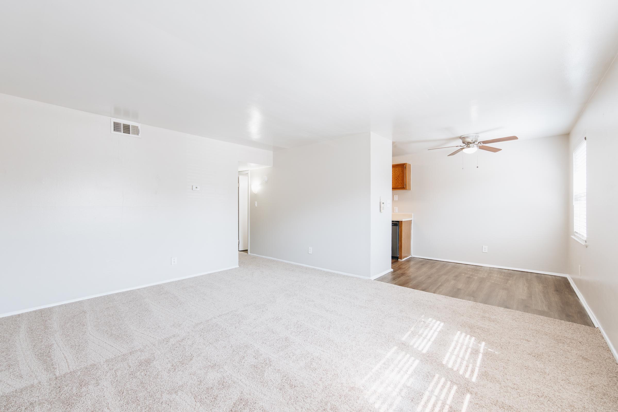 Empty living room with light-colored carpet and white walls. A ceiling fan is mounted in the center, and there is a small kitchen area visible to the right with wooden cabinetry. Natural light floods the space through a window, creating a bright and open atmosphere.
