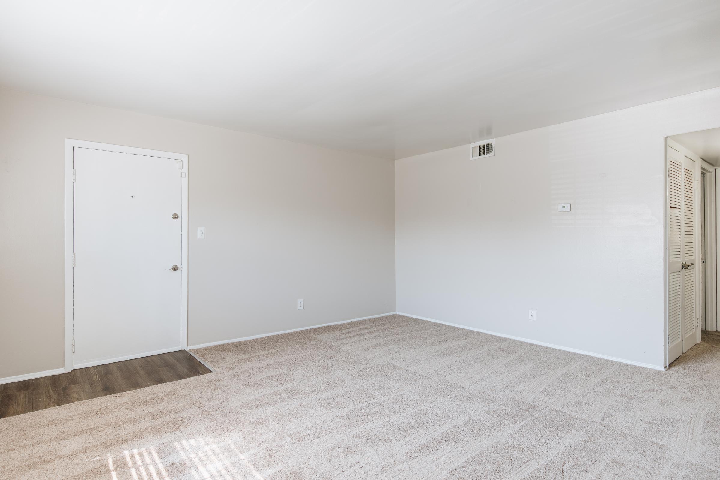 A spacious, empty room with light beige walls and a carpeted floor. There is a closed white door on the left and a small entry space with a mat. Natural light streams in from an unseen window, highlighting the clean, uncluttered space. On the right, a closet with white sliding doors is partially visible.