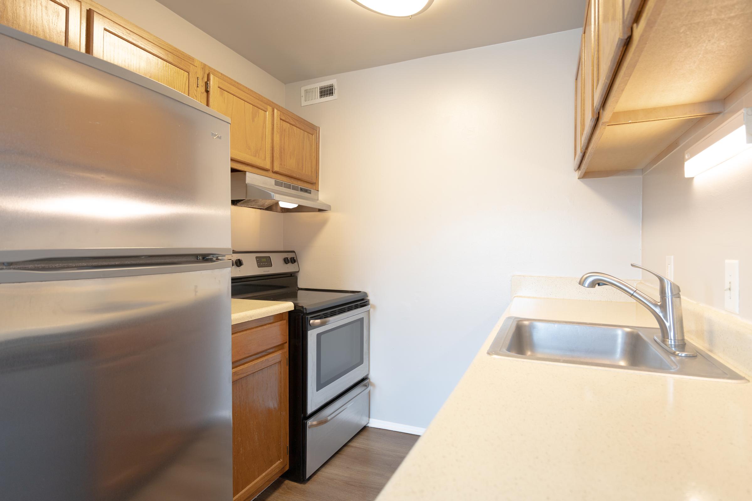 A modern kitchen featuring wooden cabinetry, stainless steel refrigerator, black oven and stovetop, and a stainless steel sink. The countertop is light-colored and the walls are painted in a neutral tone, providing a clean and spacious appearance.