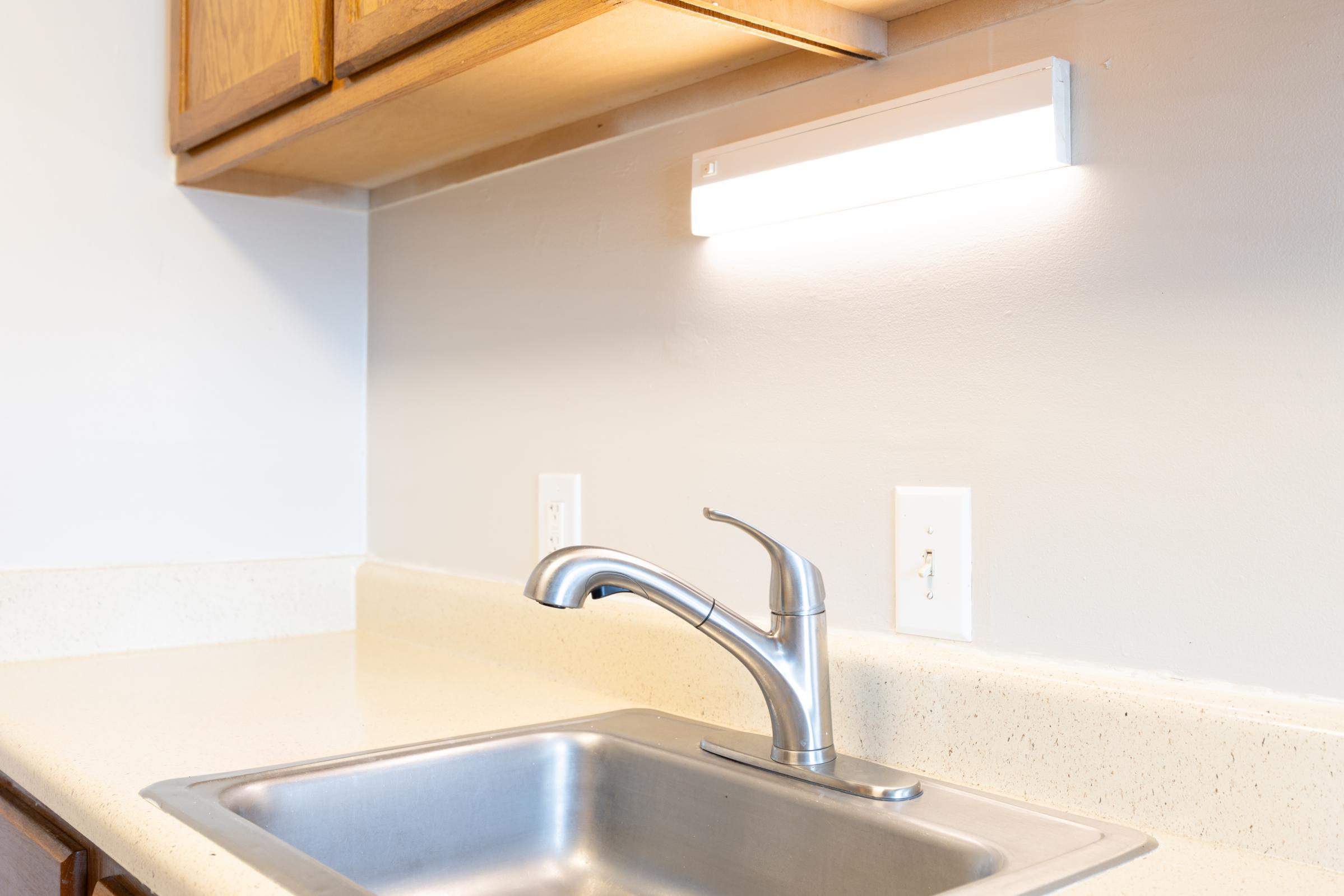 A modern kitchen sink with a shiny silver faucet, set against a light-colored countertop and wall. Above the sink, there is a sleek, minimalist light fixture illuminating the area. Wooden cabinets are visible to the left, complementing the clean and functional design of the space.