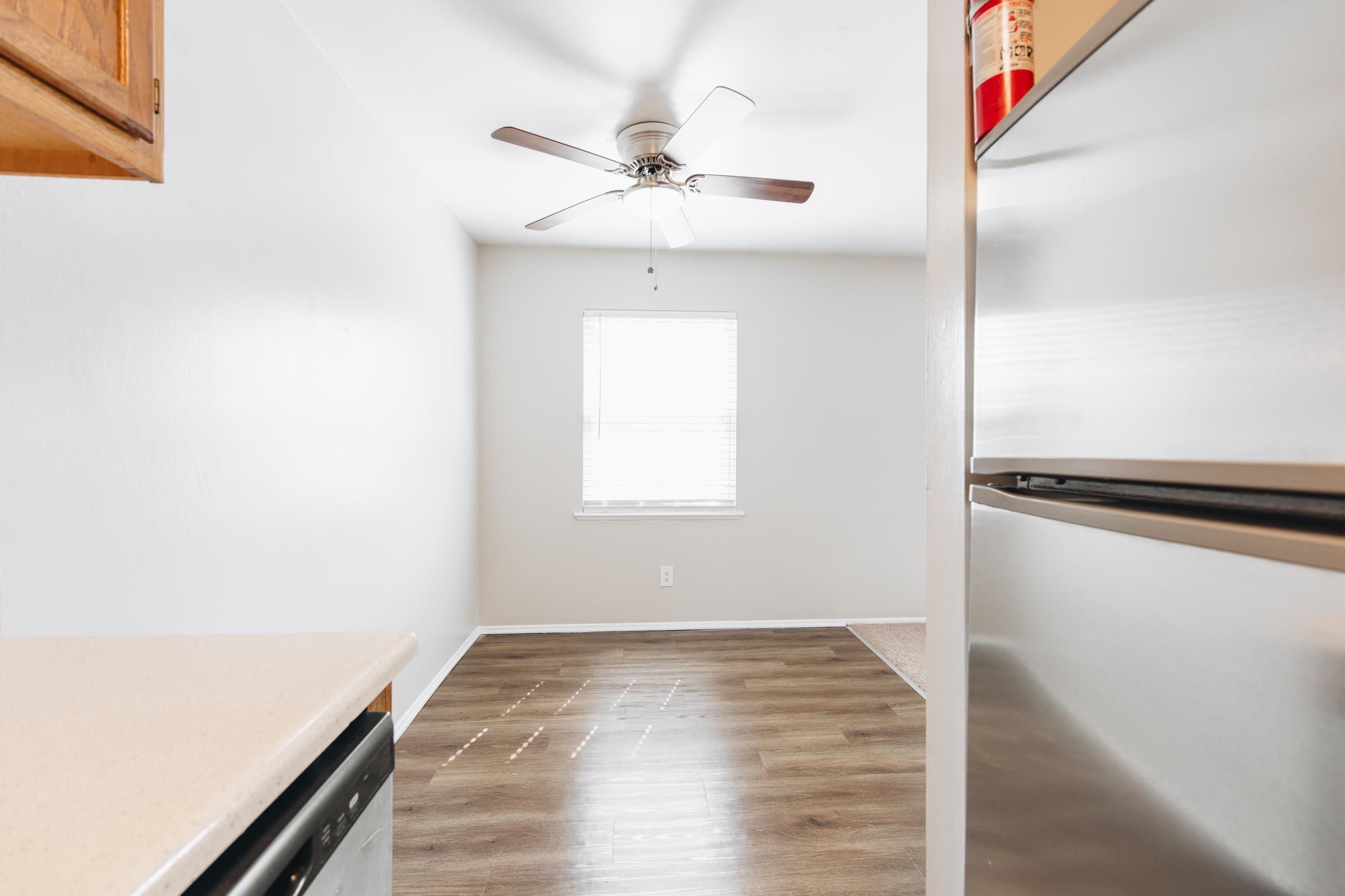 Bright and clean kitchen with light-colored walls, a ceiling fan, and a window allowing natural light. The flooring is a warm wood tone, and a countertop and stainless steel refrigerator are visible. A small area with carpet is seen in the background, creating a cozy atmosphere.