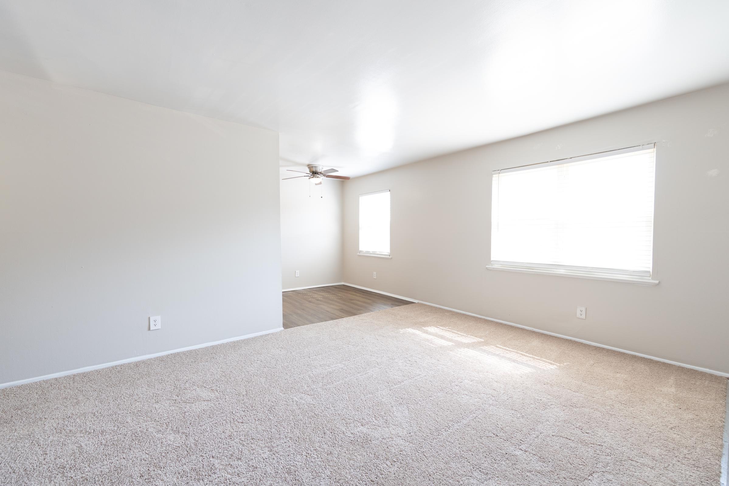 Spacious, empty living room with light-colored carpet, a ceiling fan, and large windows allowing natural light. The walls are painted a neutral color, and there is an adjoining area that leads to additional light from another window. The space is clean and ready for furnishings.