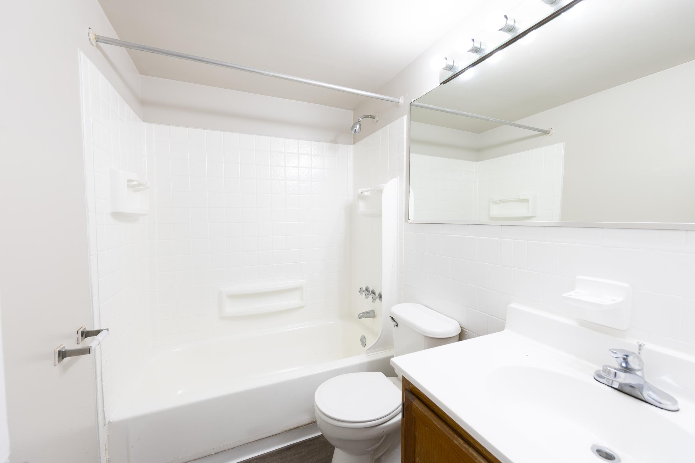 A clean and simple bathroom featuring a bathtub-shower combo, a toilet, and a single sink with a mirror above it. The walls are tiled in white, and the room has minimal decor and natural lighting.