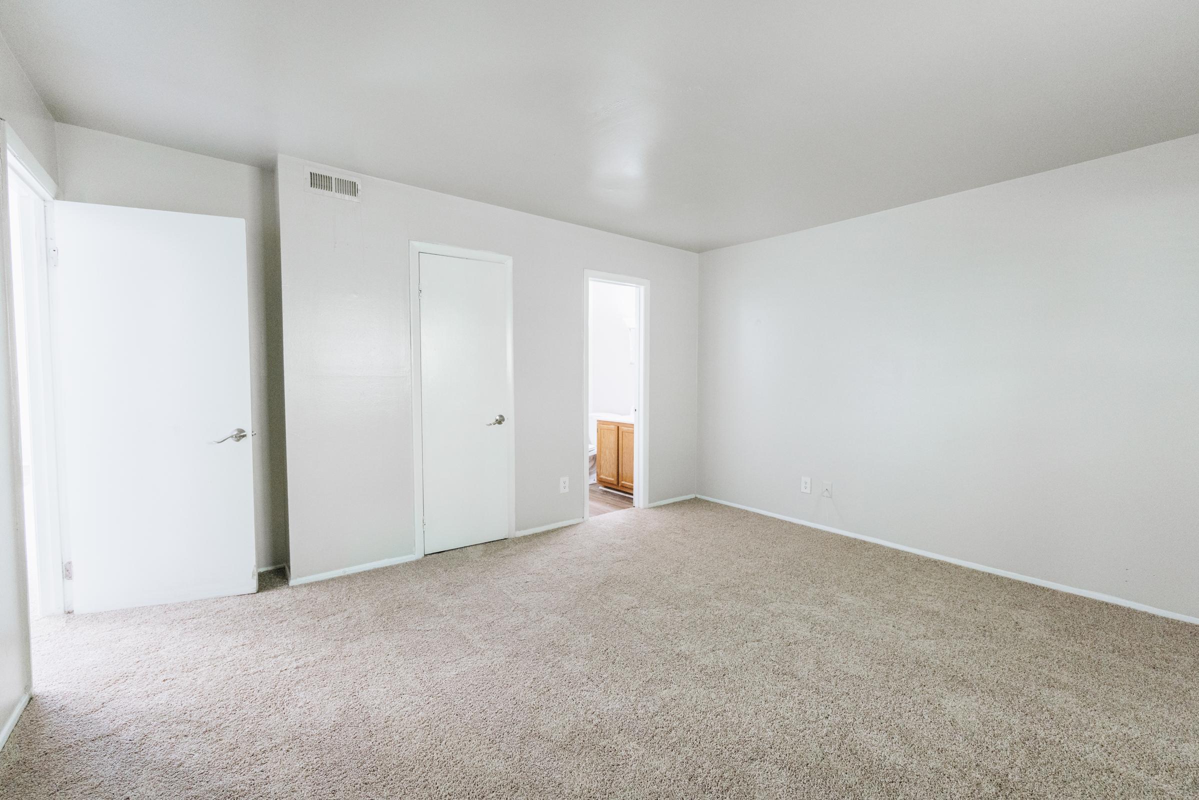 A spacious, empty room with light-colored walls and a beige carpet. Two doors are visible: one leading to a bathroom and the other an entry or closet. Natural light illuminates the room, creating an airy feel. No furniture is present, emphasizing the room’s size and potential for different layouts.