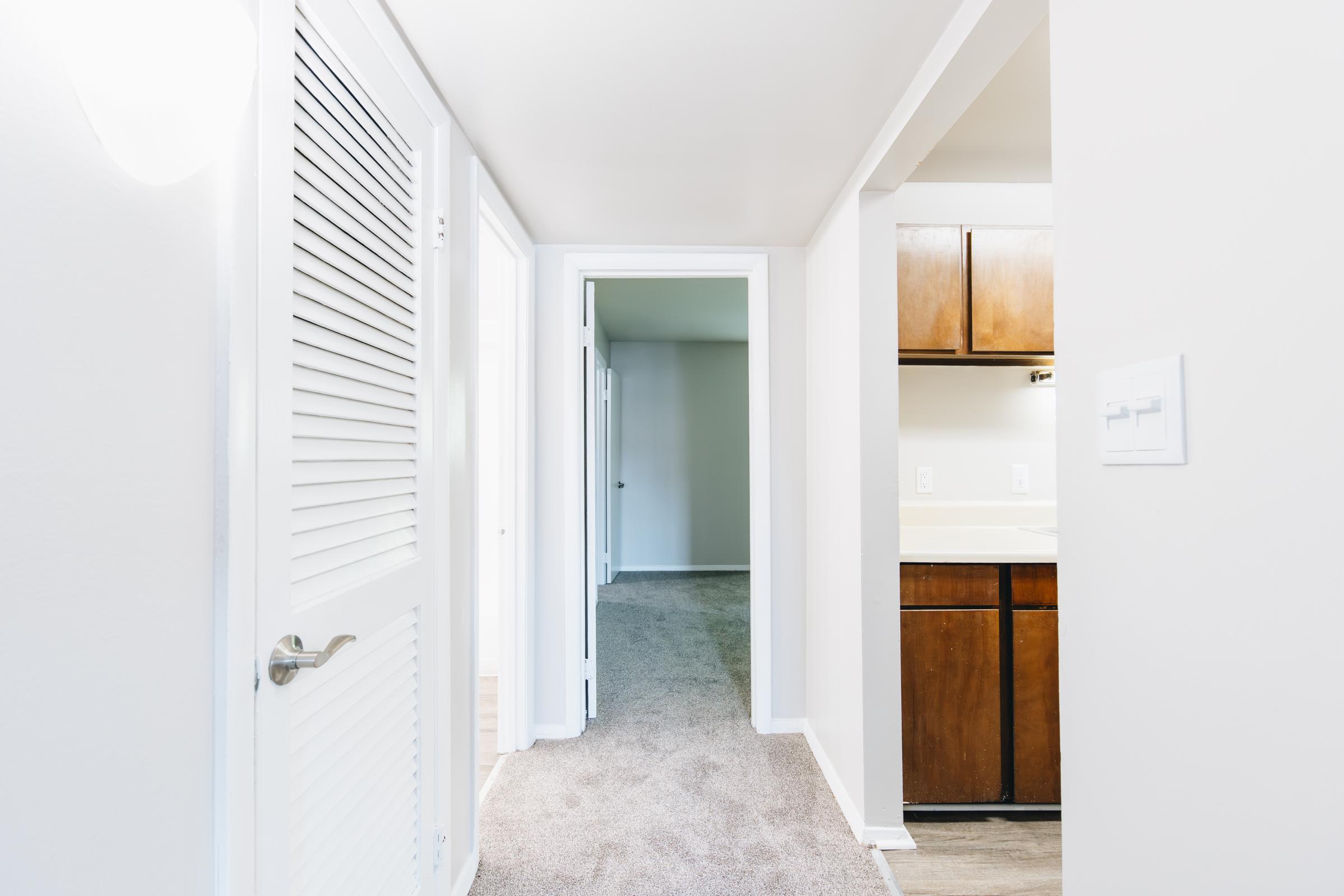 A well-lit hallway in an interior space features white walls and carpeted floors. On the left, a door with horizontal slats opens to another room, while ahead, a doorway leads to a spacious area. To the right, brown cabinets are visible in a kitchen or utility area, completing a clean and modern aesthetic.