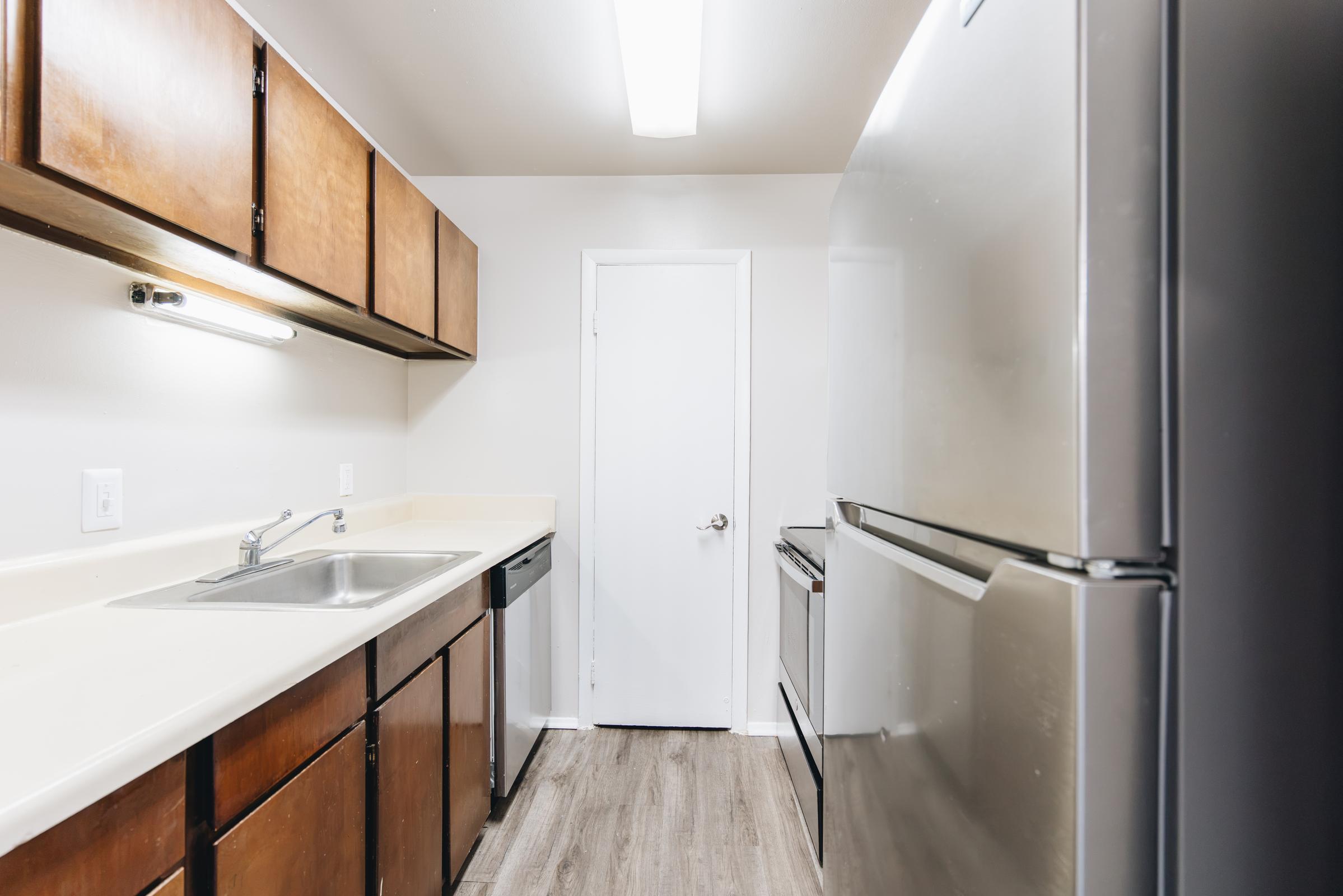 A view of a modern kitchen with wooden cabinets, a stainless steel refrigerator, a sink, and a dishwasher. The walls are painted white, and there is a door at the end of the room. The flooring is a light wood laminate. Overall, the kitchen appears clean and well-lit with overhead lighting.
