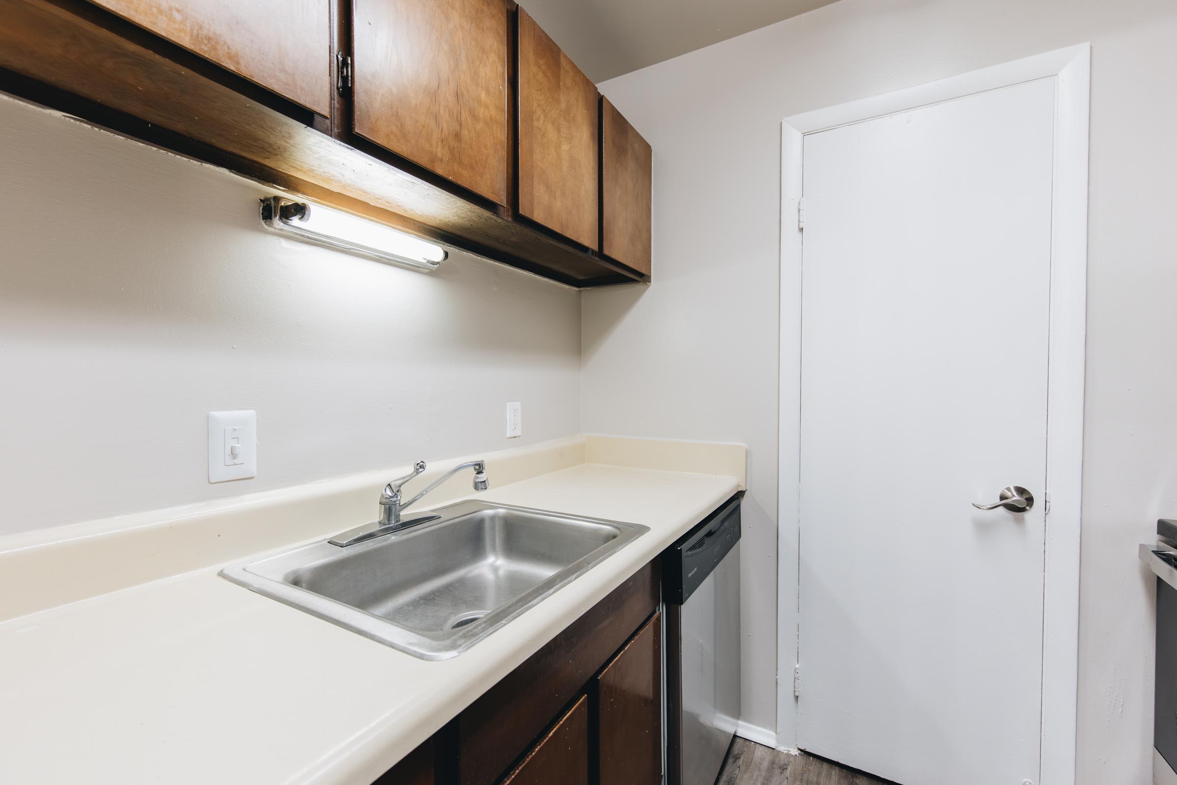 A small kitchen area featuring a light brown cabinet above a white countertop with a stainless steel sink. The kitchen has a white door leading to another room and a dark dishwasher. The walls are painted a light color, enhancing the overall brightness of the space.