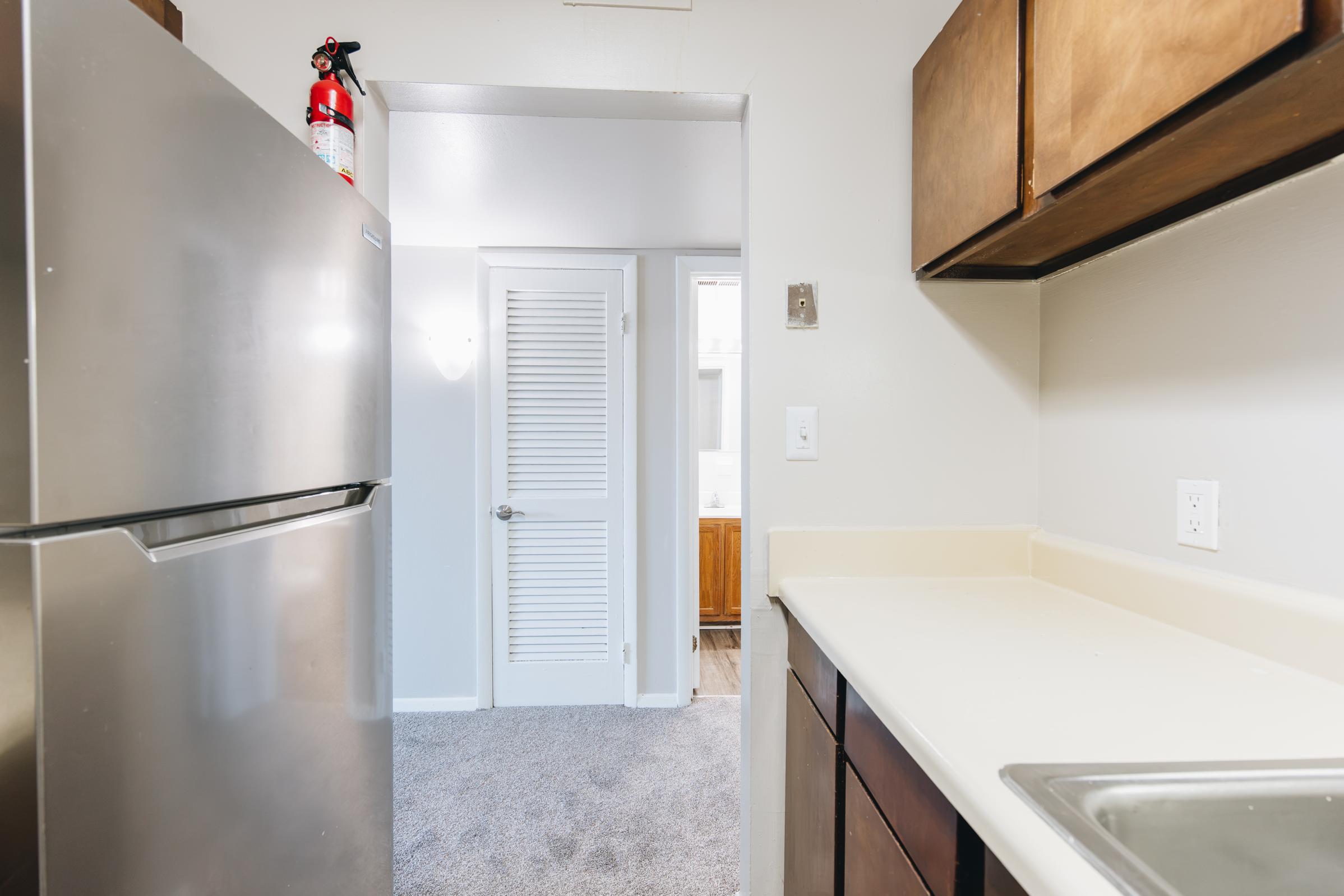 A view of a kitchen with a stainless steel refrigerator and a sink. The countertop is beige, and there are wooden cabinets above. In the background, a door leads to another room, and there is a fire extinguisher mounted on the wall. The floor is carpeted, creating a cozy atmosphere.