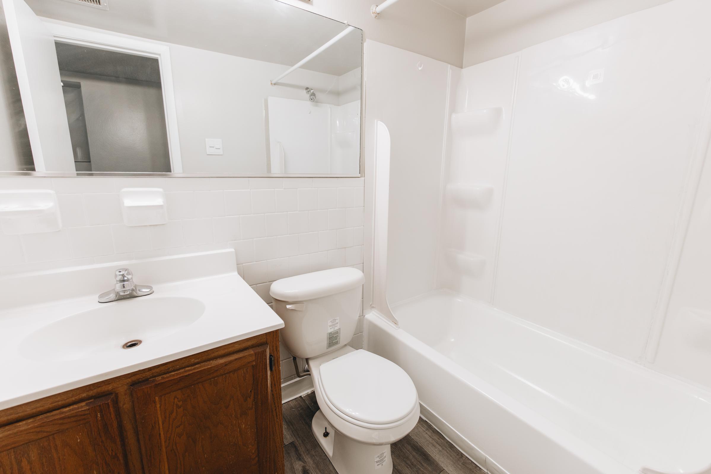 A clean and modern bathroom featuring a white bathtub and shower combination, a toilet, and a vanity with a sink. The walls are tiled white, and there is a large mirror above the sink. The flooring is wood-like, creating a warm atmosphere in the space.