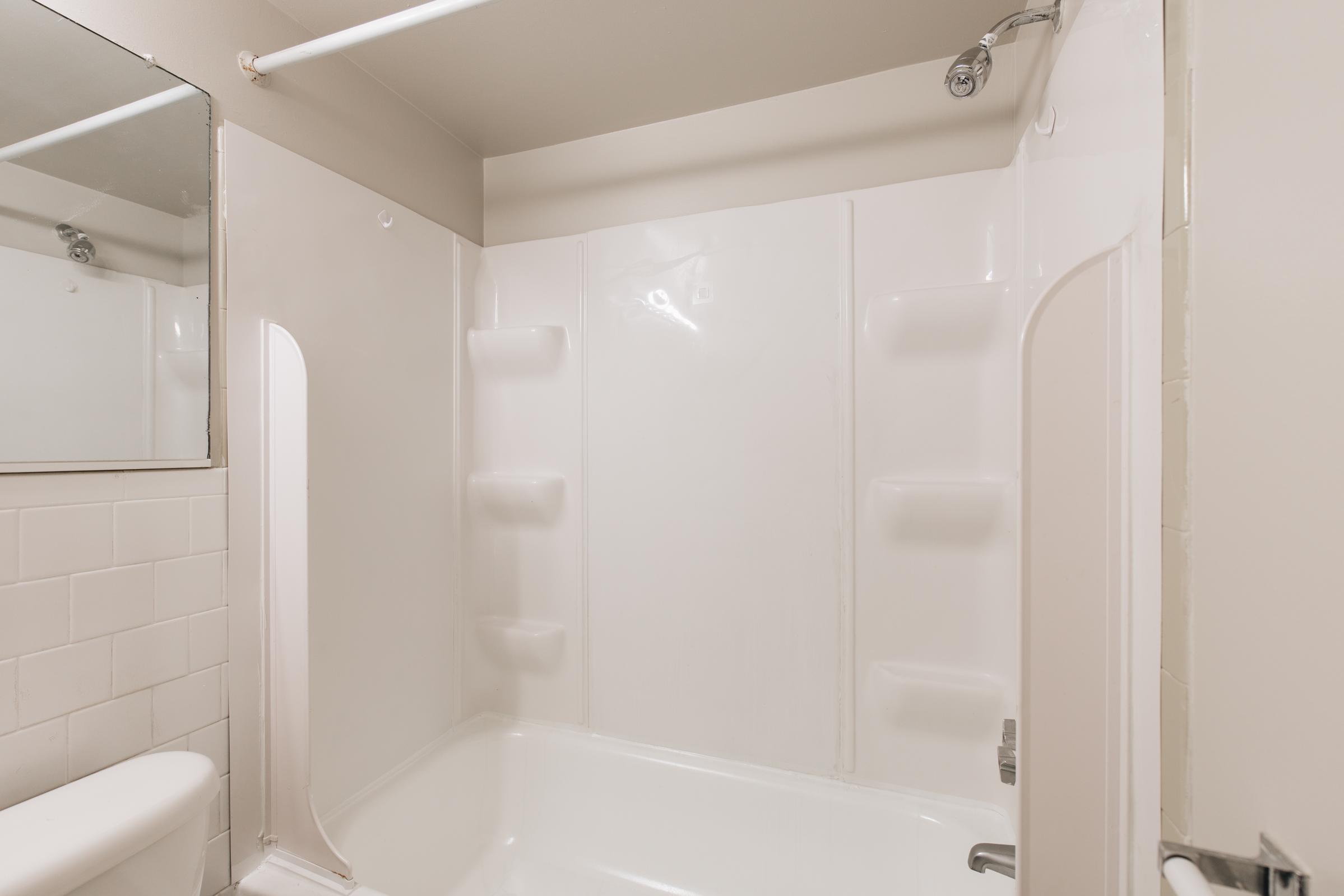 A clean, modern bathroom featuring a white shower stall with a sliding glass door. The walls are light-colored with a large mirror above a white sink. The shower has built-in shelves for toiletries, and there’s a white bathtub below the shower area. Natural light illuminates the space.