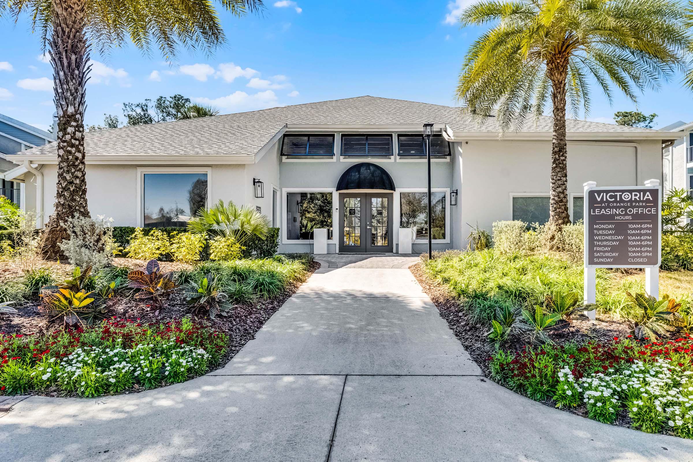 A modern leasing office building surrounded by lush greenery and colorful flowers. Palm trees line the pathway leading to the entrance, which features large windows and an arched doorway. A sign indicates it's the Victoria Leasing Office. Bright blue sky in the background.