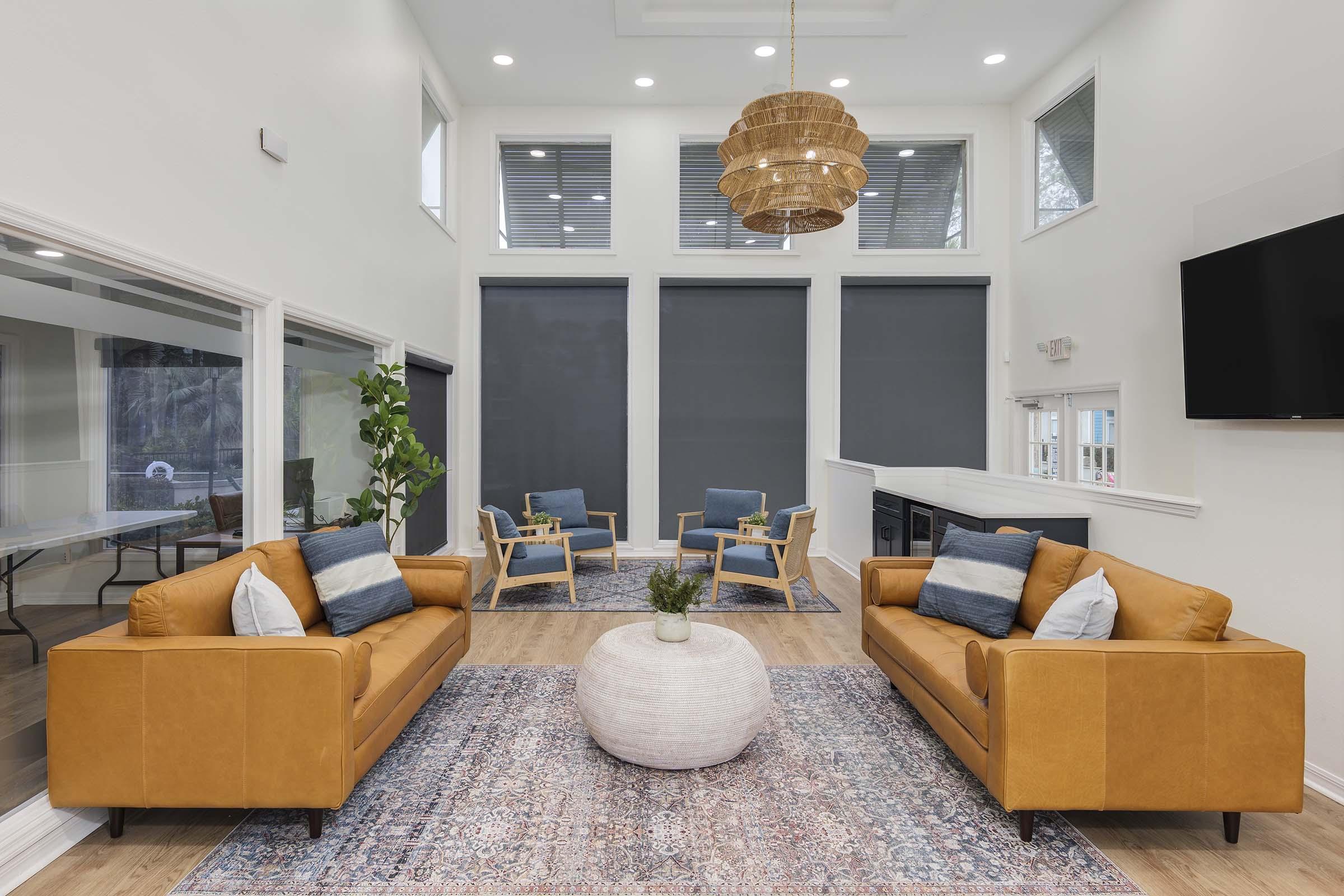 A modern living room featuring two tan leather sofas facing each other, a round white coffee table in the center, and a neutral-toned area rug. Light-filled space with high ceilings, large windows dressed with gray shades, and stylish blue accent chairs in the background. A decorative pendant light hangs above.