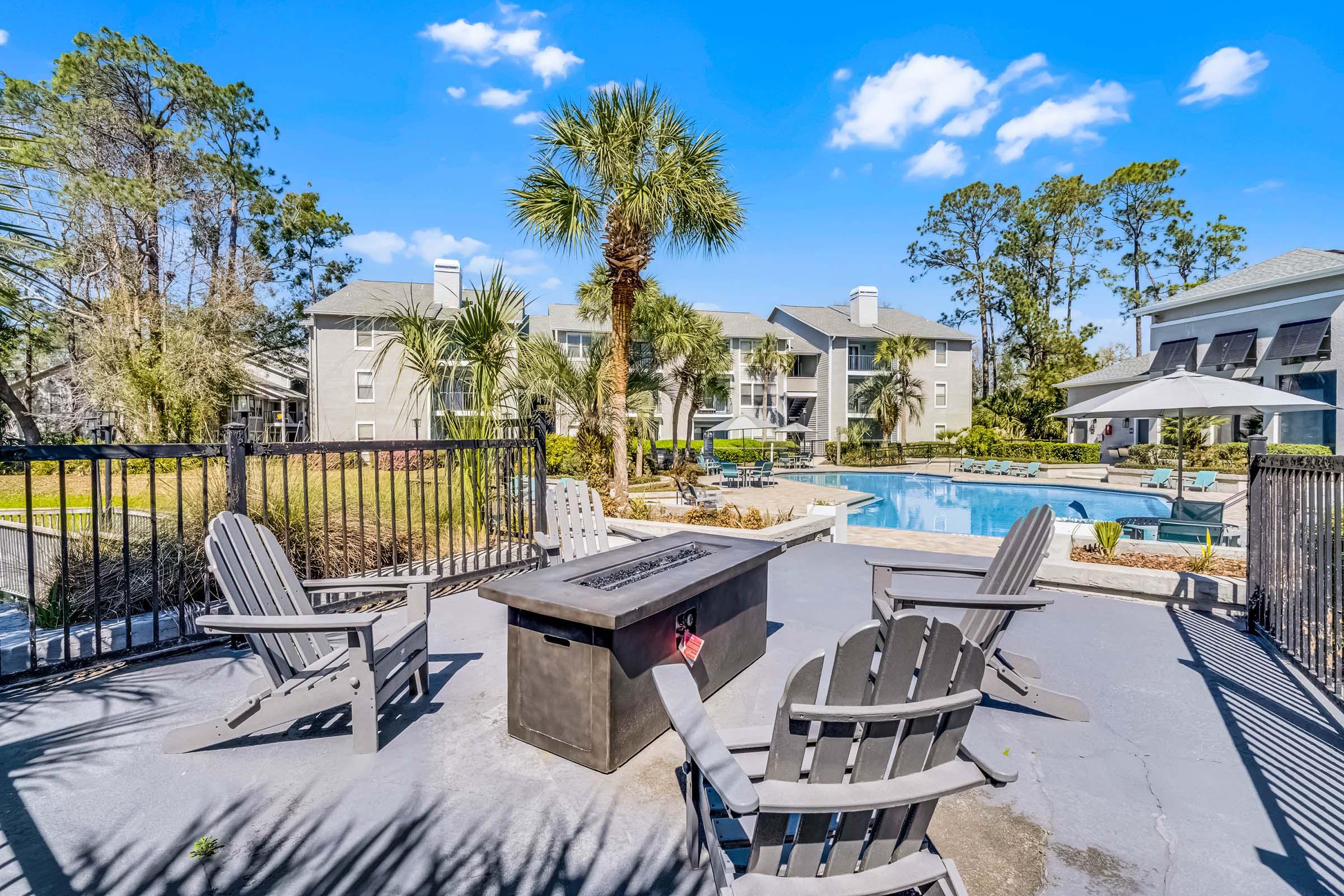 A sunny outdoor area featuring a pool surrounded by palm trees and greenery. In the foreground, there are several Adirondack chairs arranged around a fire pit, with an apartment complex visible in the background. Bright blue sky and fluffy white clouds enhance the inviting atmosphere.