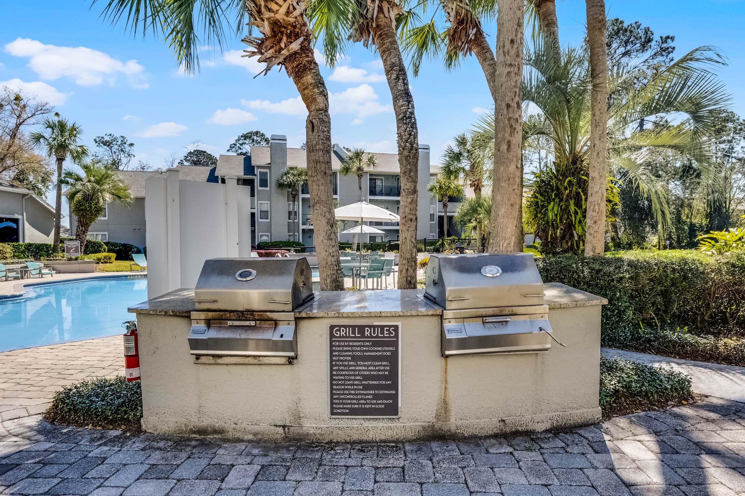 A barbecue grill area featuring two grills, surrounded by palm trees and lush greenery. In front of the grills, there is a sign outlining "Grill Rules." The background includes a swimming pool area with lounge chairs and umbrellas, creating a relaxing outdoor setting.