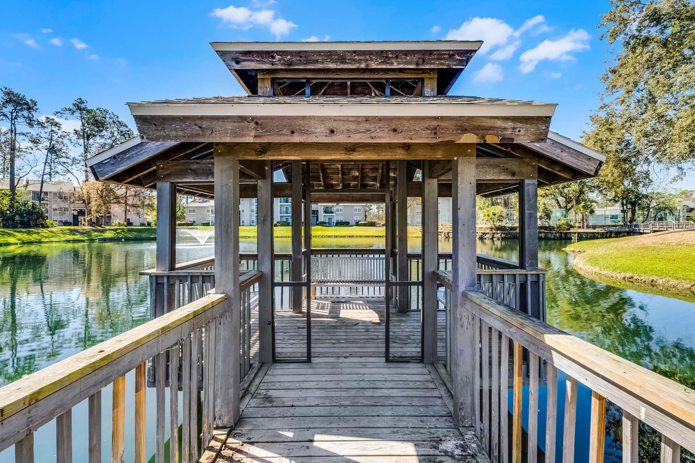 A wooden gazebo on a calm lake, featuring a deck that extends over the water. The structure has a sloped roof and open sides, surrounded by lush green grass and trees. In the background, there are buildings visible along the shore. The sky is clear with a few fluffy clouds.