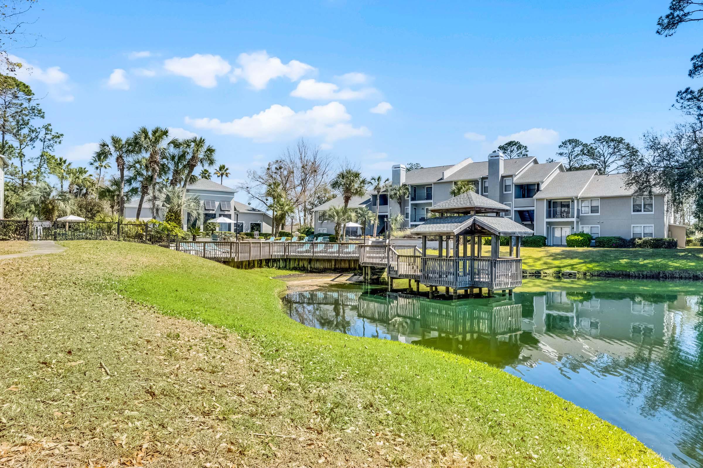 A tranquil scene featuring a pond surrounded by lush greenery and palm trees. In the background, there's a residential complex with gray buildings. A wooden gazebo extends over the water, and a bridge connects to the path leading to the complex, creating a serene outdoor setting.