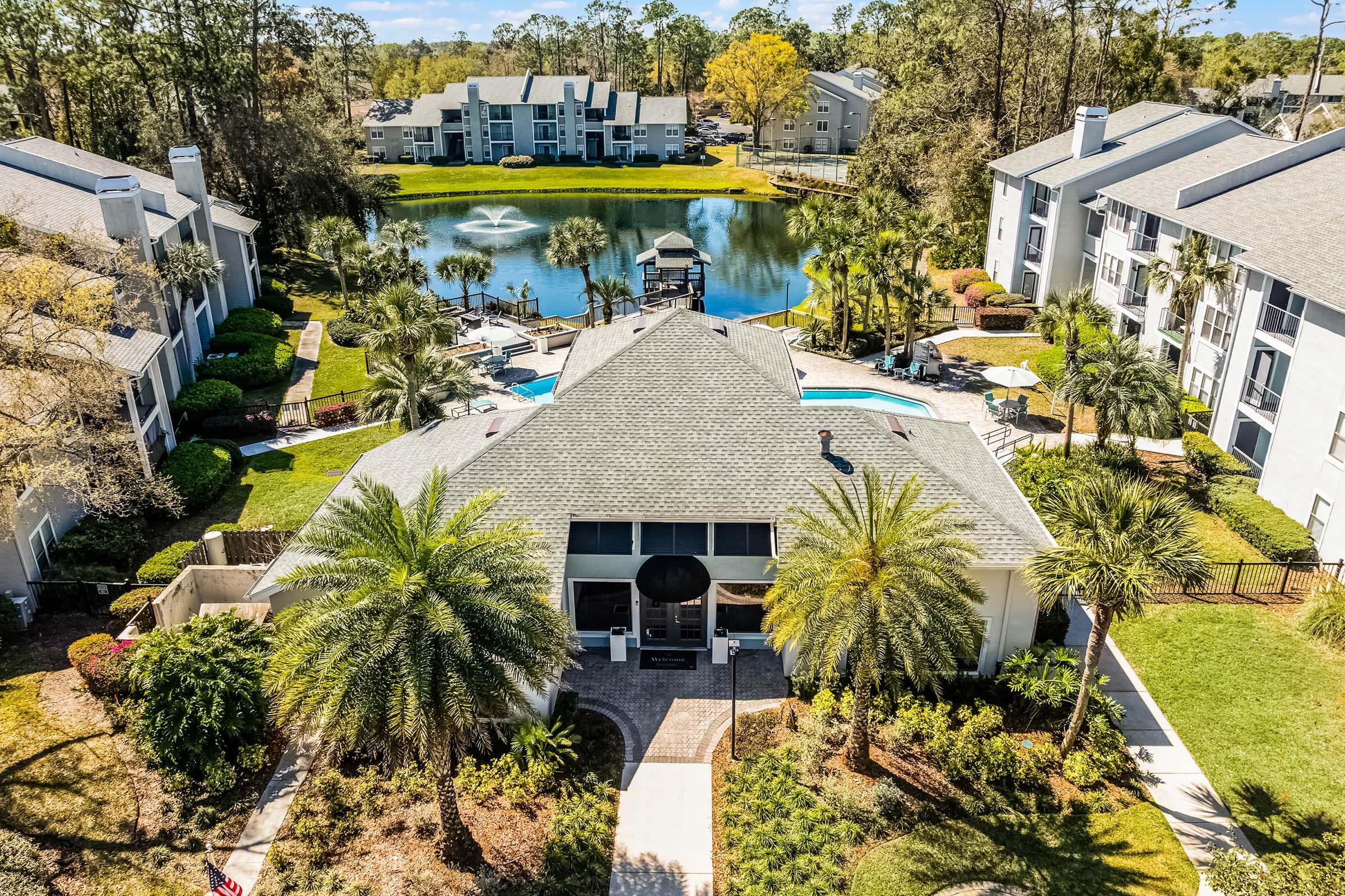 Aerial view of a residential complex featuring a central building surrounded by landscaped gardens, palm trees, and a swimming pool. In the background, there is a serene pond with a fountain and several apartment buildings lining its shore. The scene is bright and sunny, showcasing a peaceful community atmosphere.