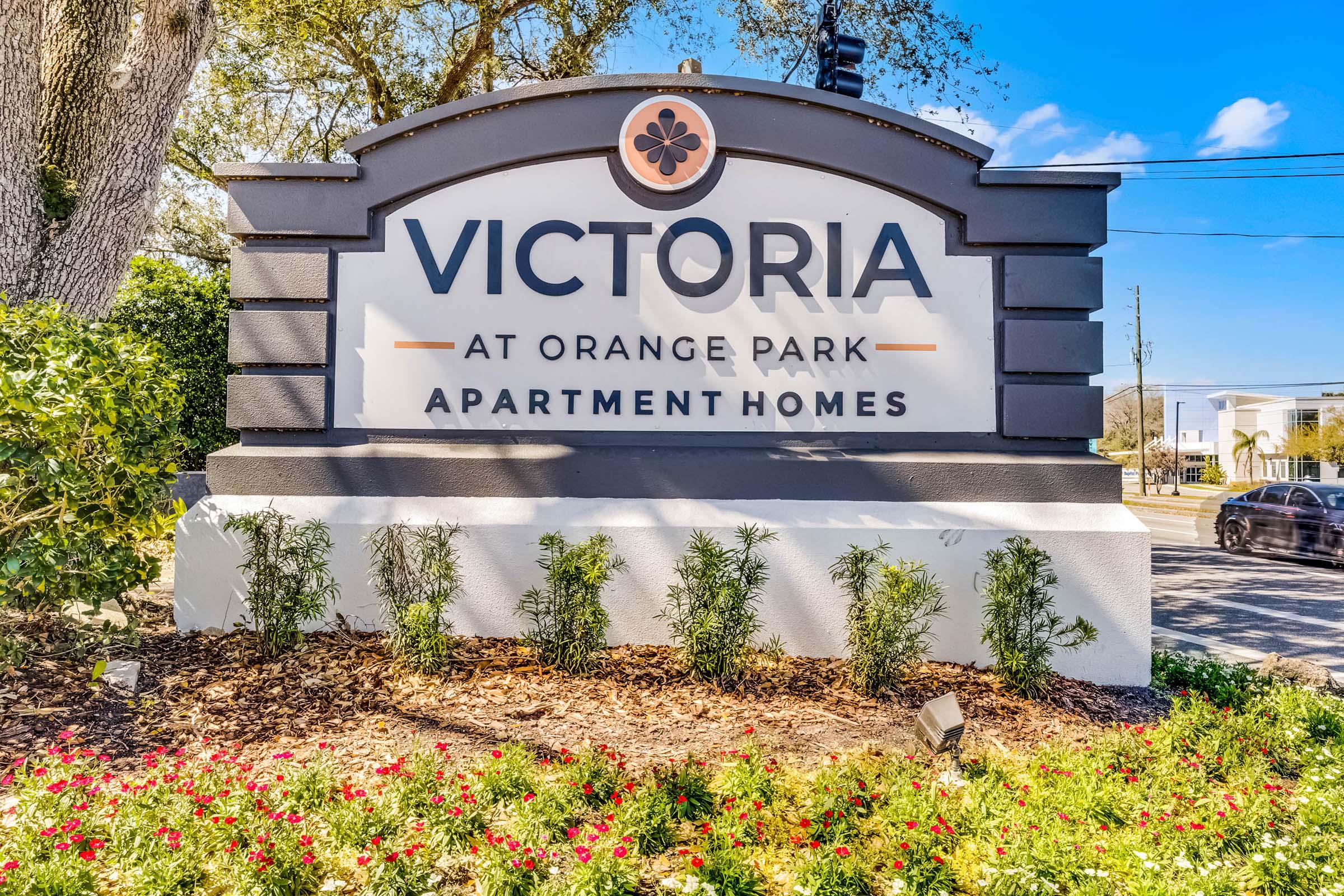 Sign for Victoria at Orange Park Apartment Homes, featuring the name "Victoria" prominently displayed with decorative elements. Surrounding the sign are well-maintained flower beds and greenery under a clear blue sky.