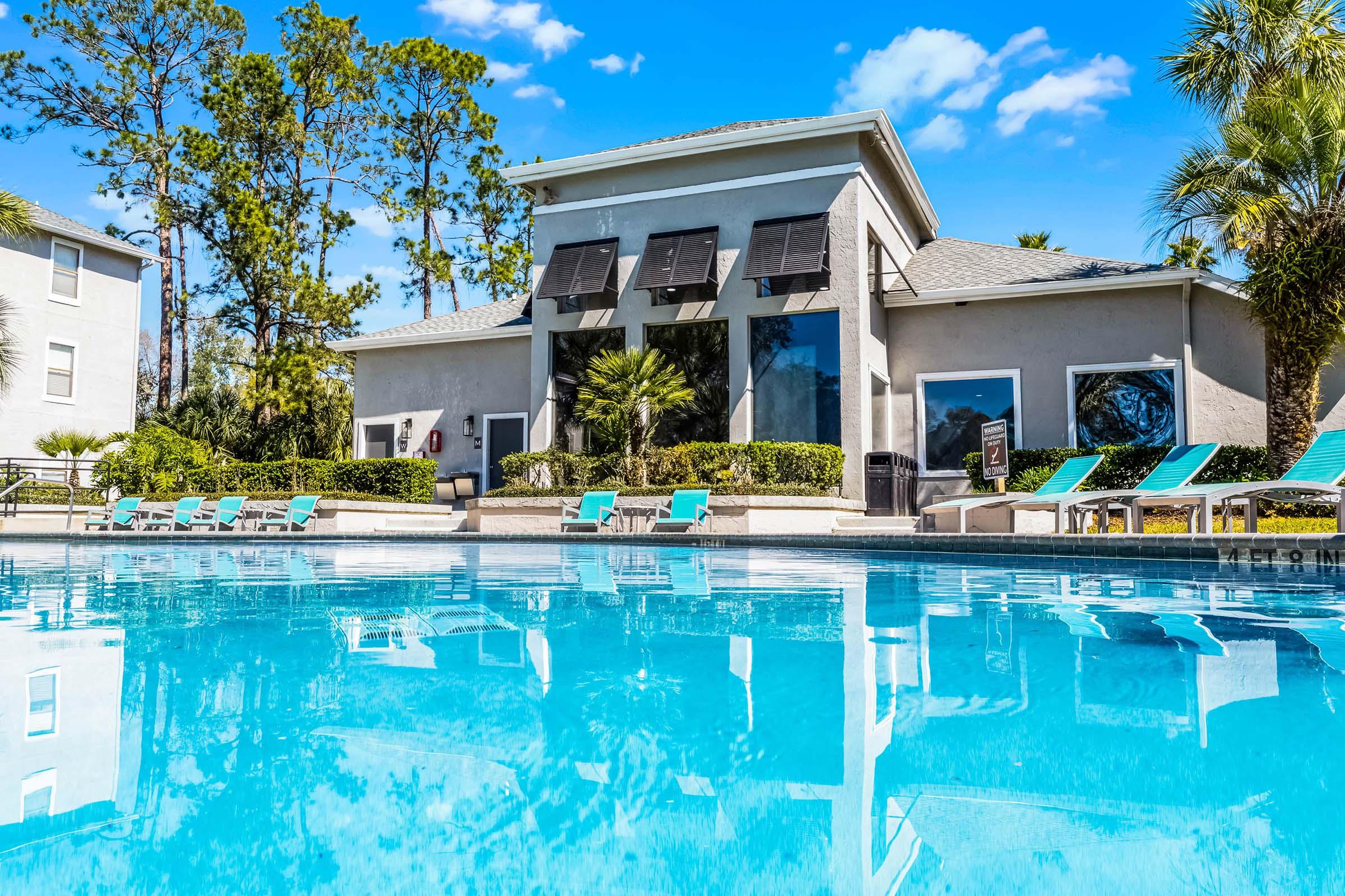 A clear blue swimming pool in the foreground with a modern building featuring large windows in the background. The building has palm trees and landscaping around it. Bright blue skies with a few clouds complete the sunny scene. Poolside loungers are arranged neatly along the edge.