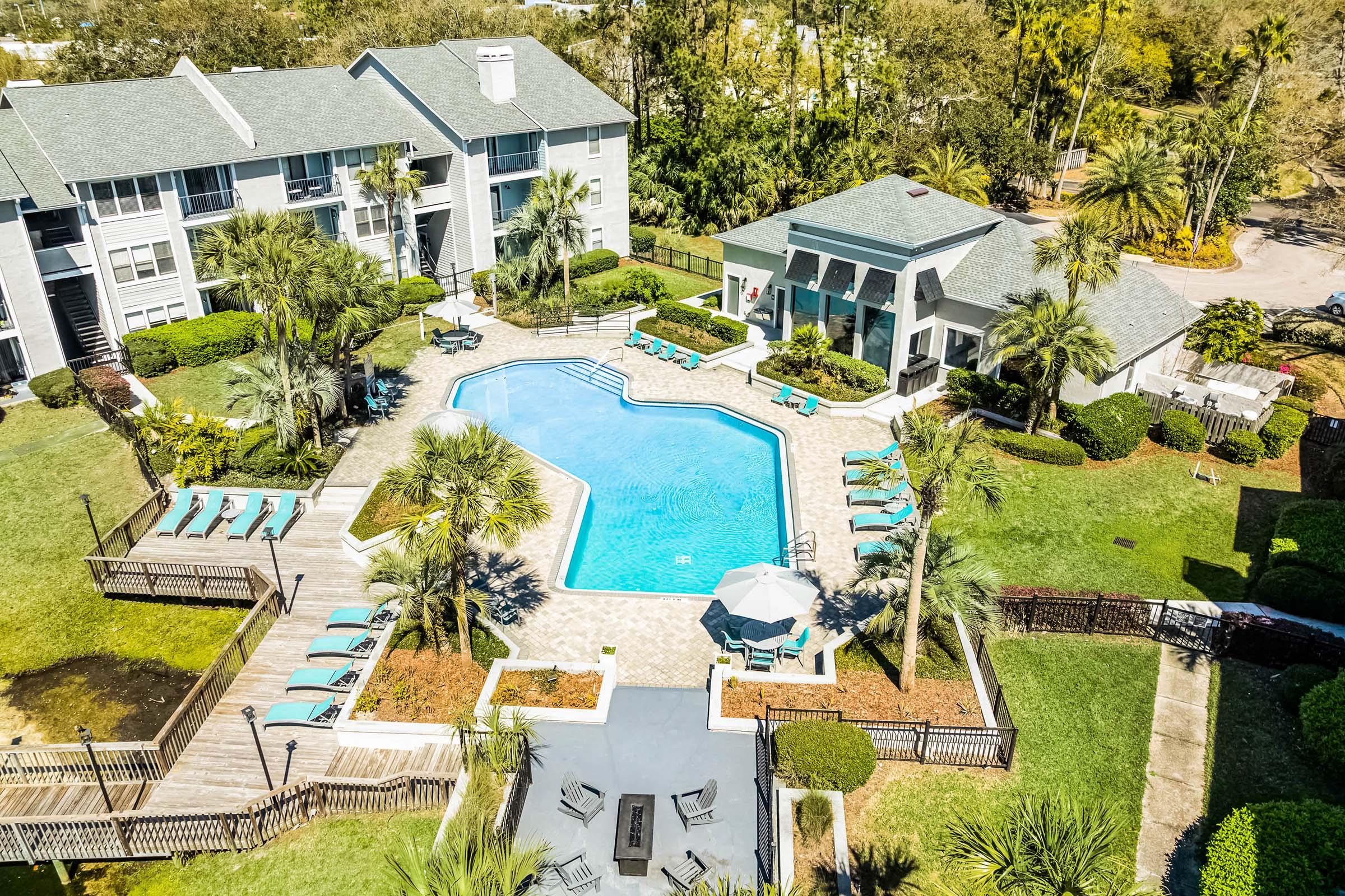 Aerial view of a landscaped pool area surrounded by residential buildings. The pool features a unique geometric shape, with lounge chairs and umbrellas arranged around it. Palm trees and manicured greenery enhance the relaxing atmosphere, and a shaded pavilion is situated near the poolside.